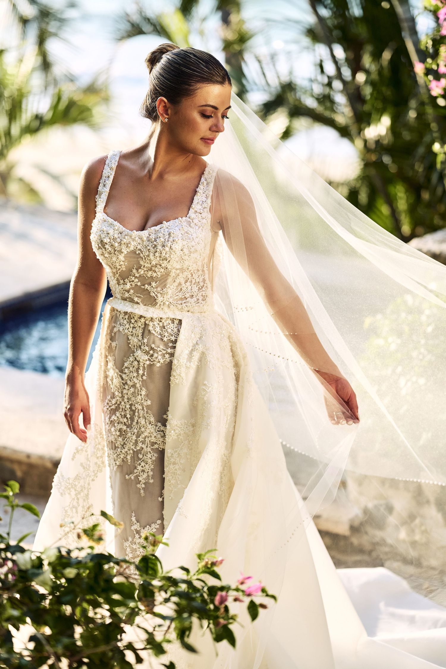 Bride in detailed lace wedding dress with flowing veil against tropical garden backdrop.