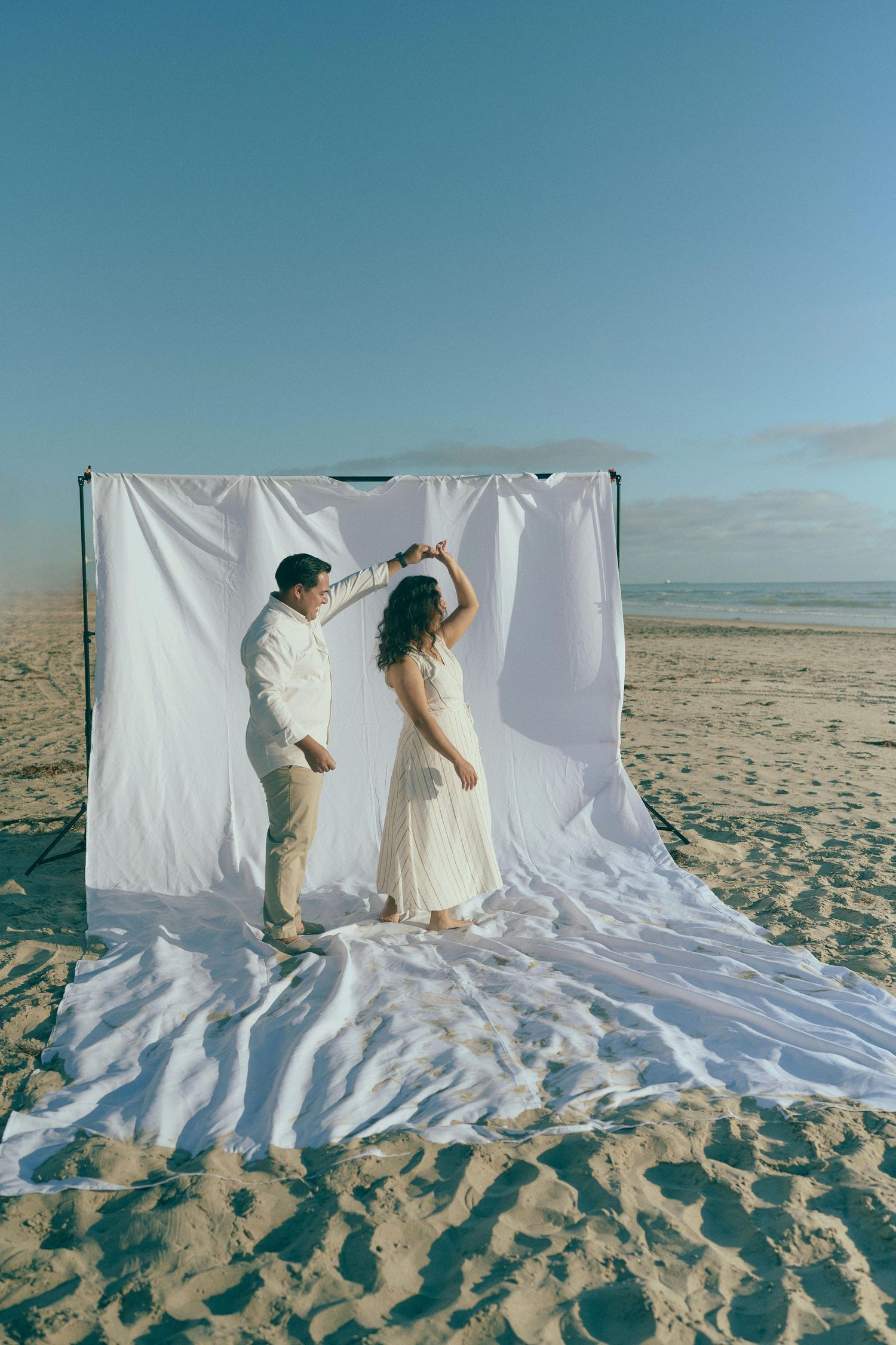 Creative beach wedding photoshoot with white fabric backdrop on sandy shores.