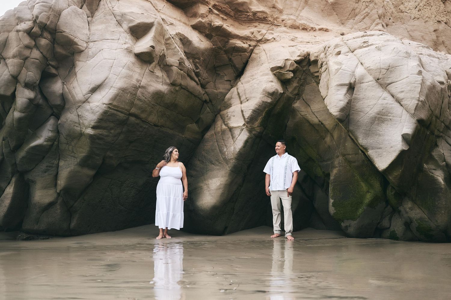 Couple in white outfits standing apart in front of dramatic rock cave formations.
