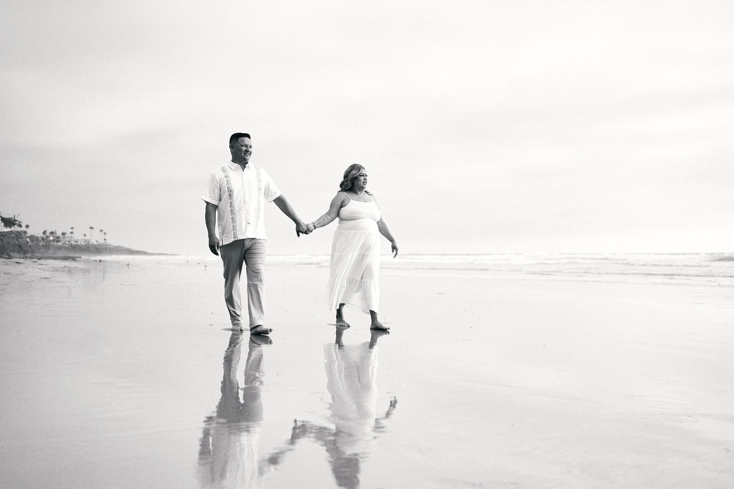 Black and white beach photo of couple holding hands while walking on wet sand.