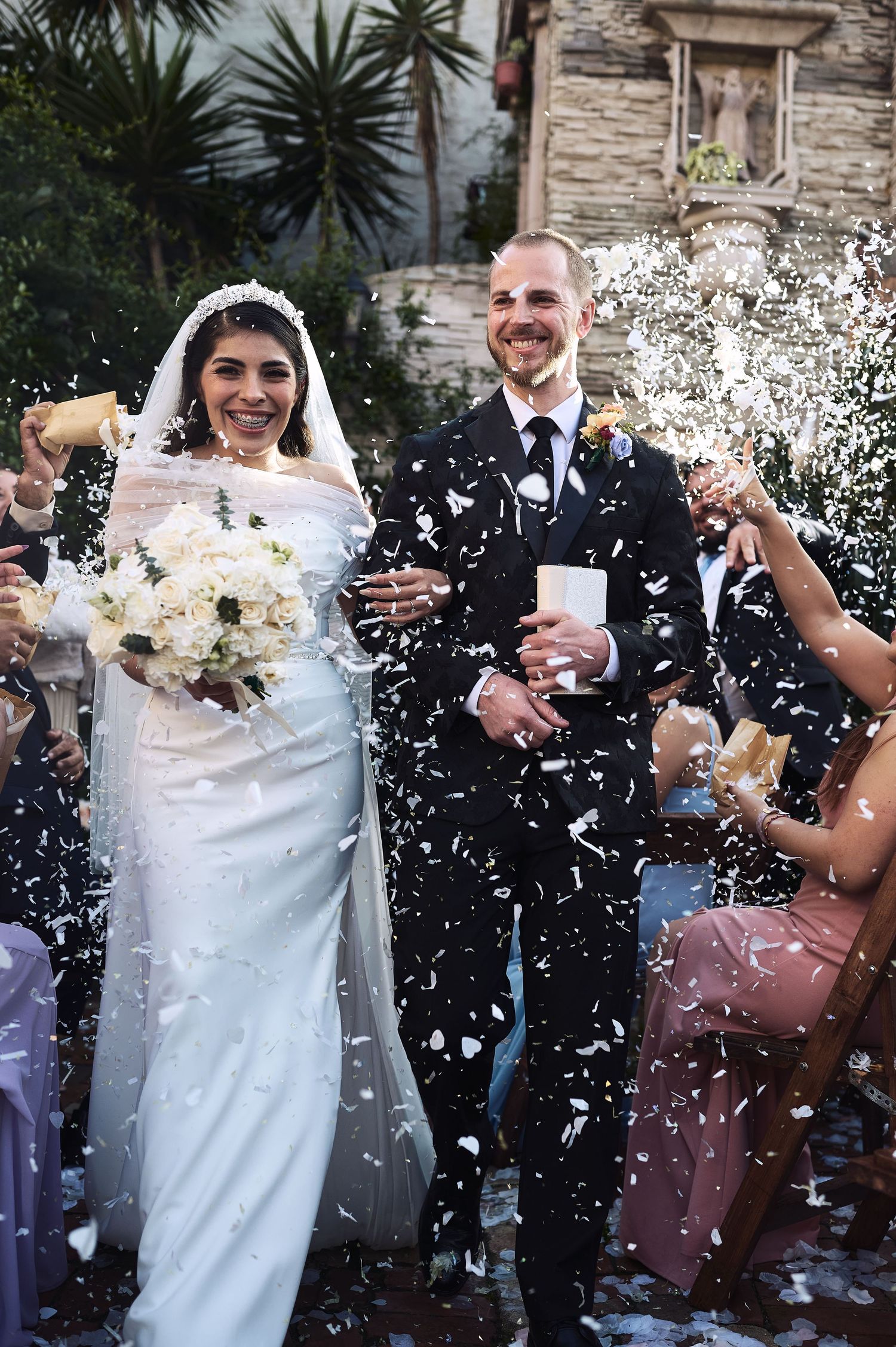 Newlyweds celebrate amid a shower of white confetti outside a stone building.