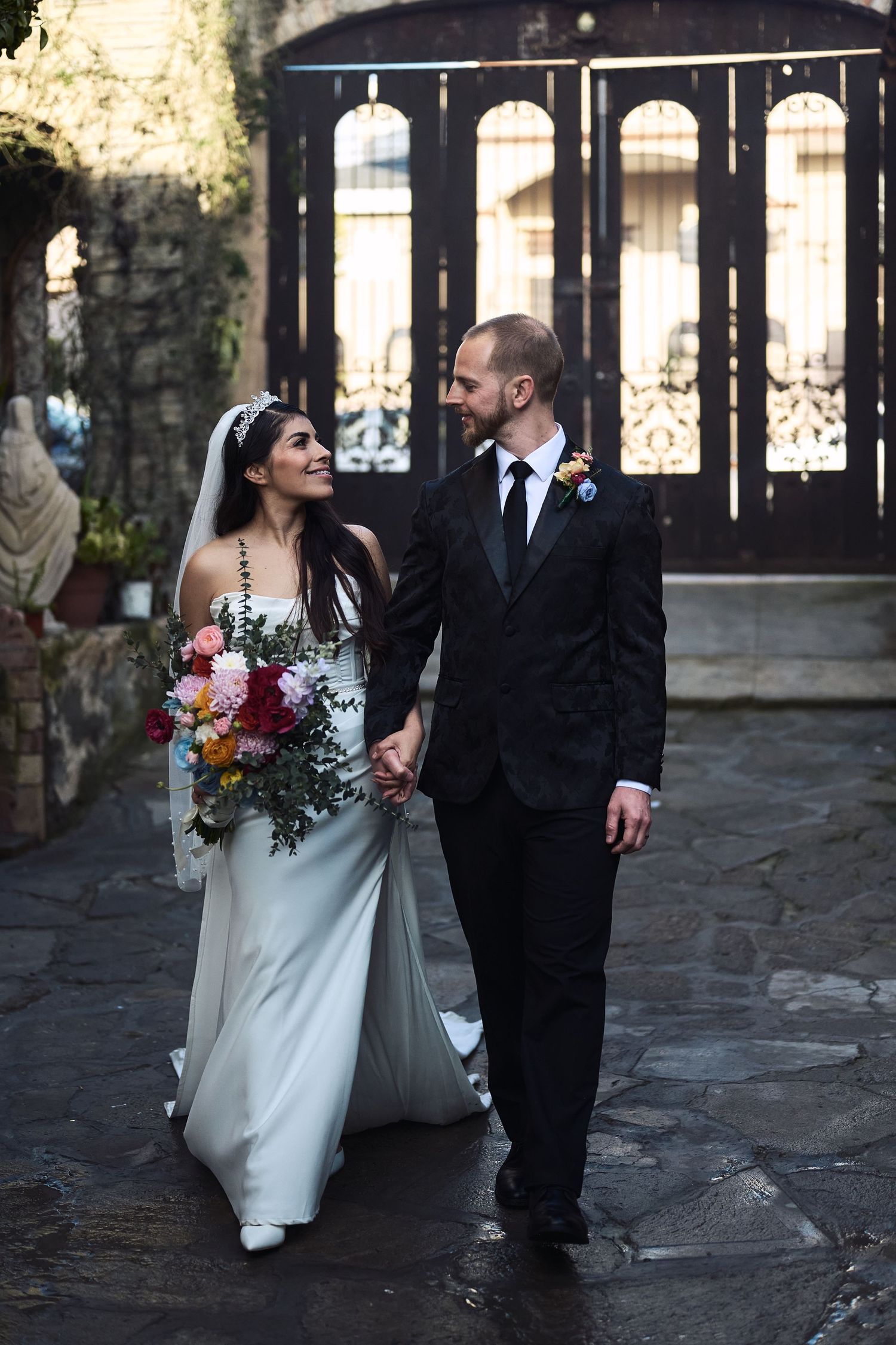 Wedding couple walking through stone archways holding colorful floral bouquets.