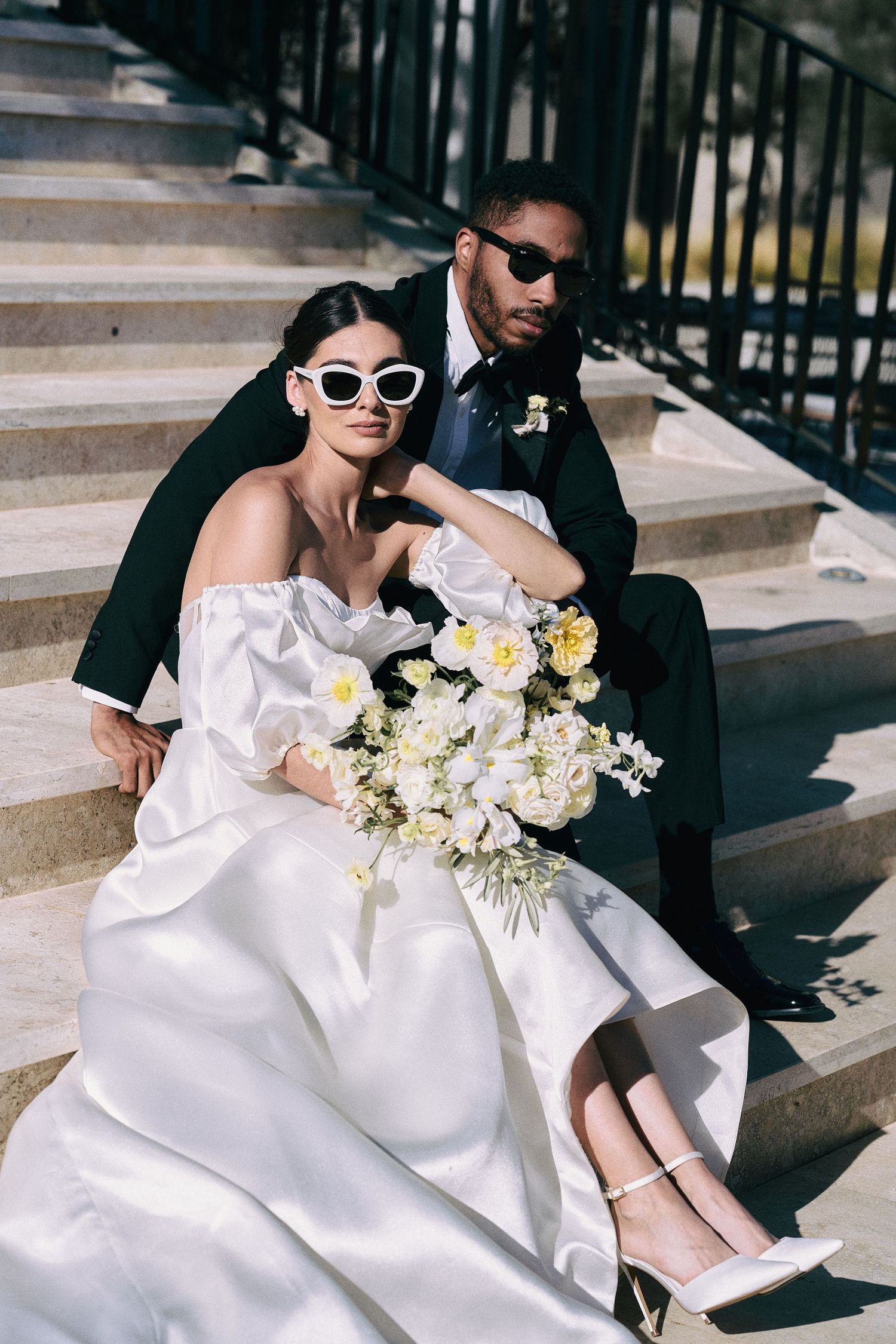 Stylish wedding couple in sunglasses posing on stone steps with white floral bouquet.