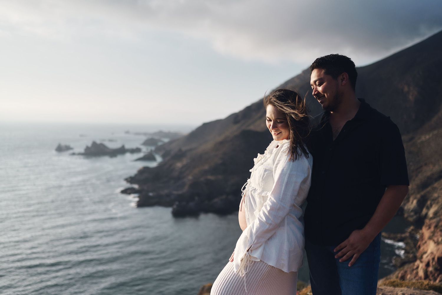 Couple embracing on coastal cliffs overlooking the ocean during sunset.