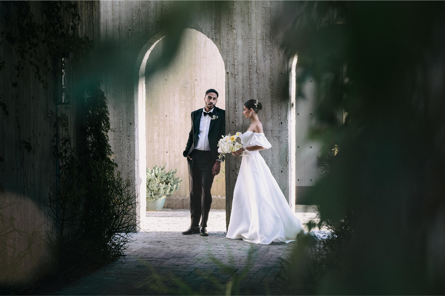A silhouetted couple in wedding attire walks through a dramatic dark corridor with bright backlighting creating an ethereal mood.