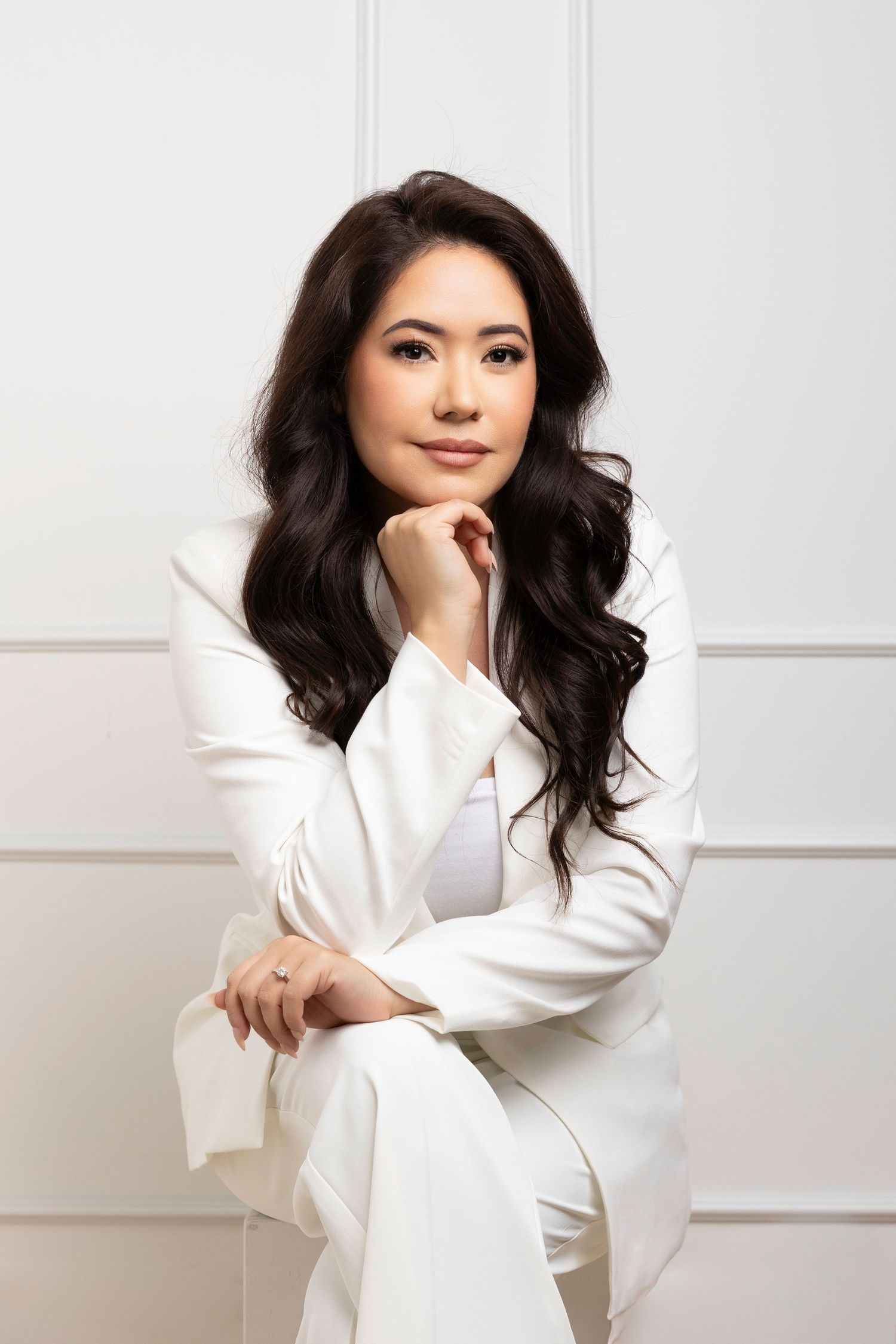 Professional portrait of someone in an elegant white pantsuit sitting with one hand under chin against a light background.