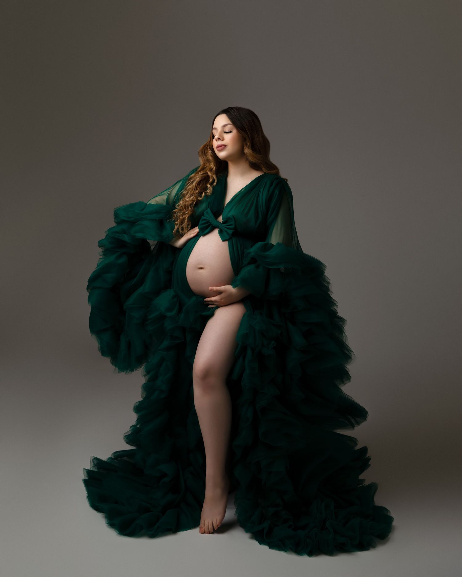 Maternity photoshoot featuring a dramatic emerald green tulle gown against a gray studio backdrop.