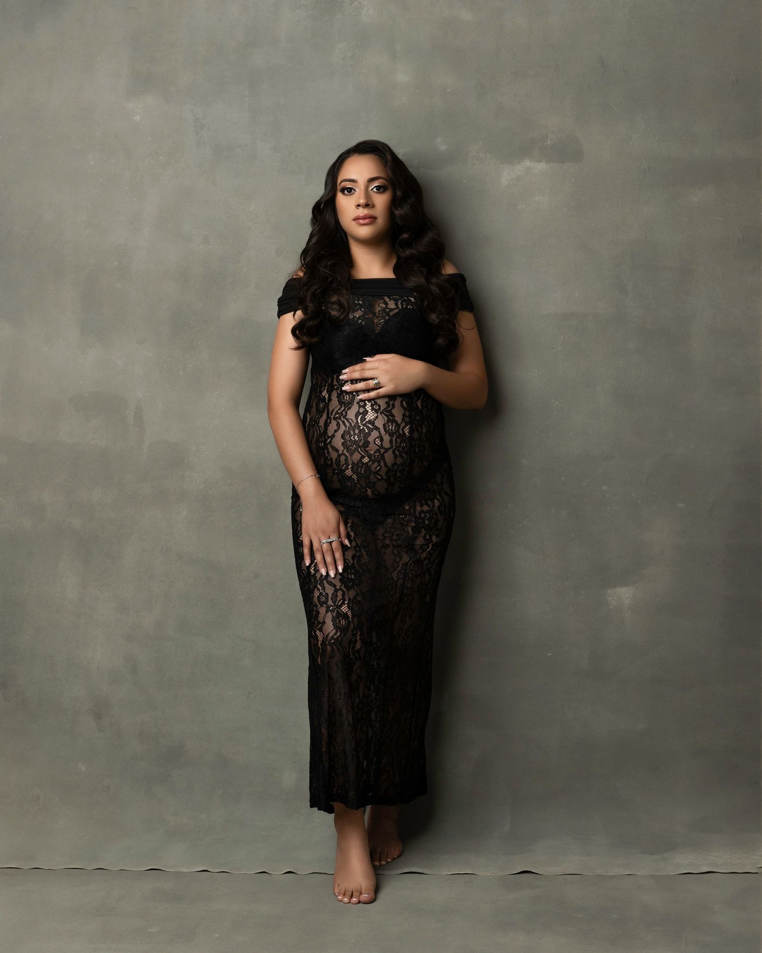 A person in a black lace dress poses elegantly against a gray textured wall in a professional studio setting.