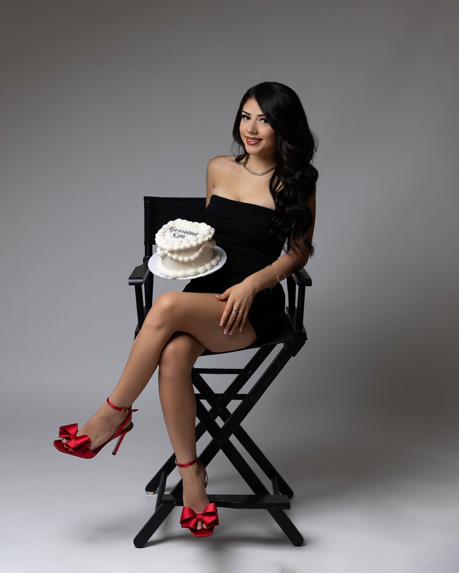 A glamour photoshoot series featuring someone in a black dress and red heels posing on a director's chair against a gray backdrop.