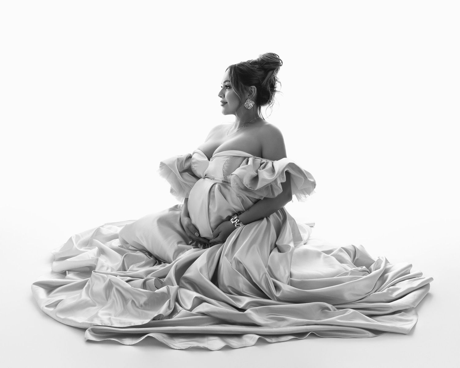 A figure in a flowing white dress sits gracefully in an artistic black and white studio portrait.