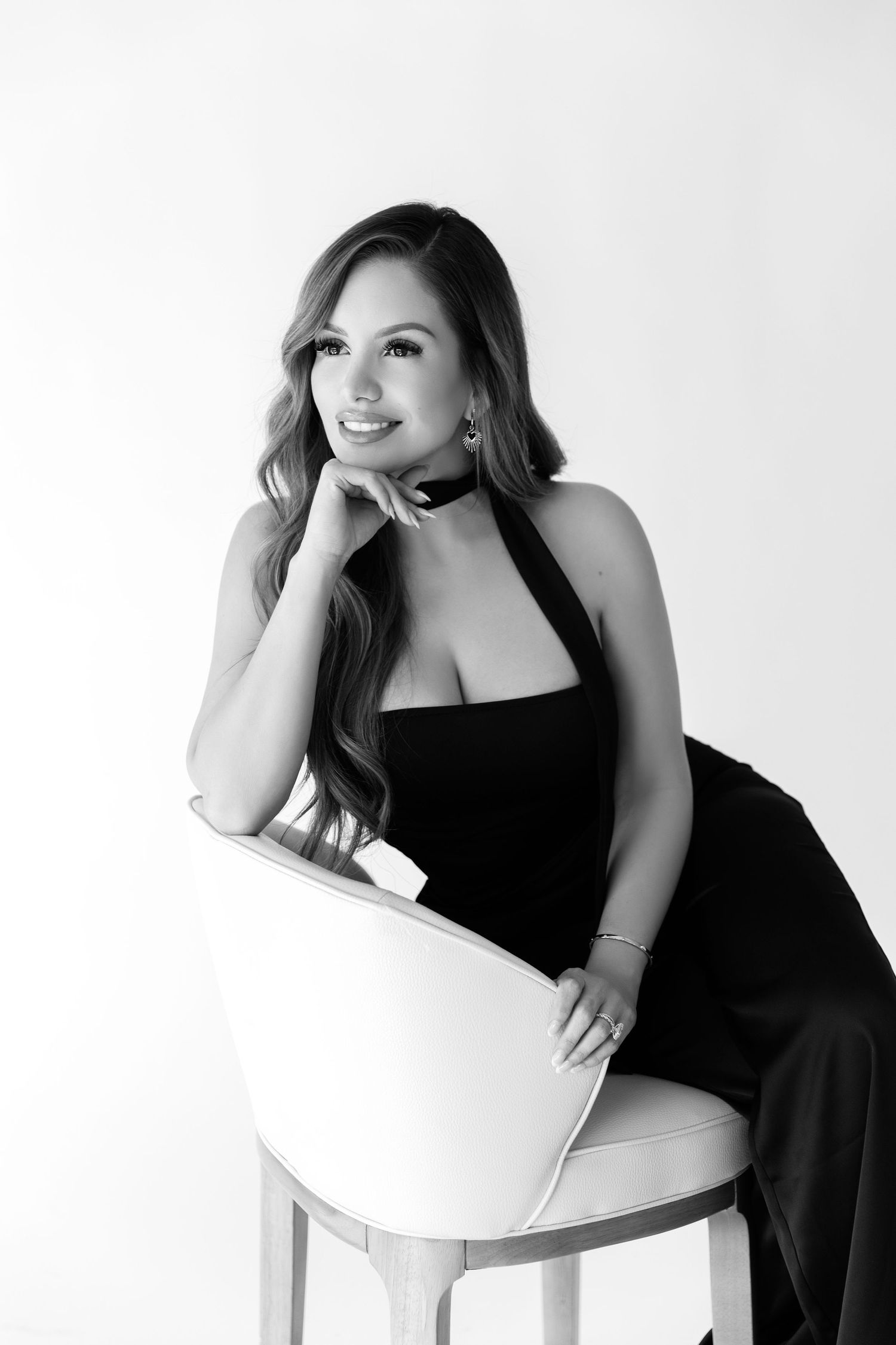 Woman in black dress poses elegantly on white chair in artistic black and white portrait series.