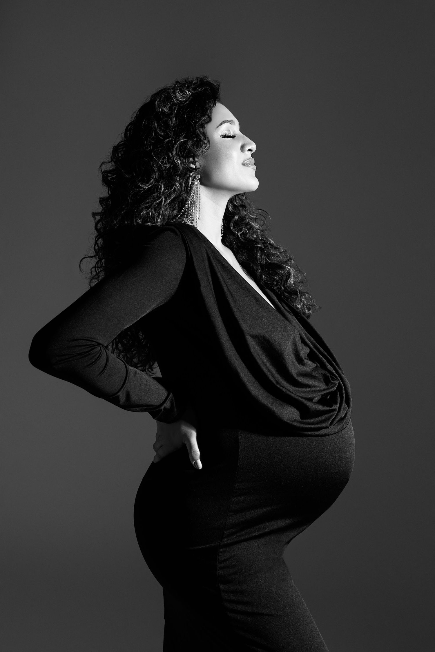 A dramatic black and white maternity portrait showing an expectant mother in a fitted black dress.