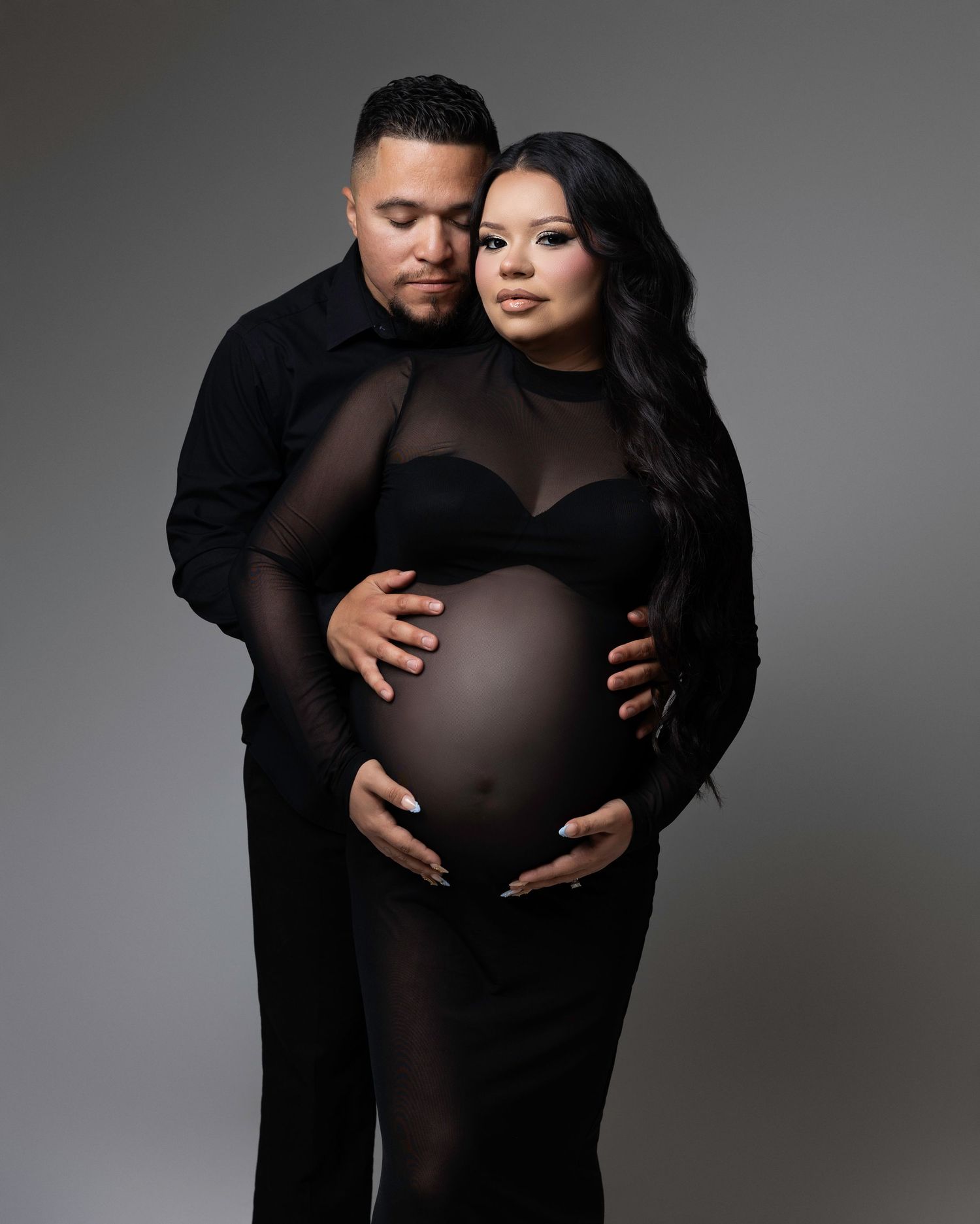 A dramatic studio portrait of an expectant couple in black attire against a grey background.  Located in Palm Desert, California.