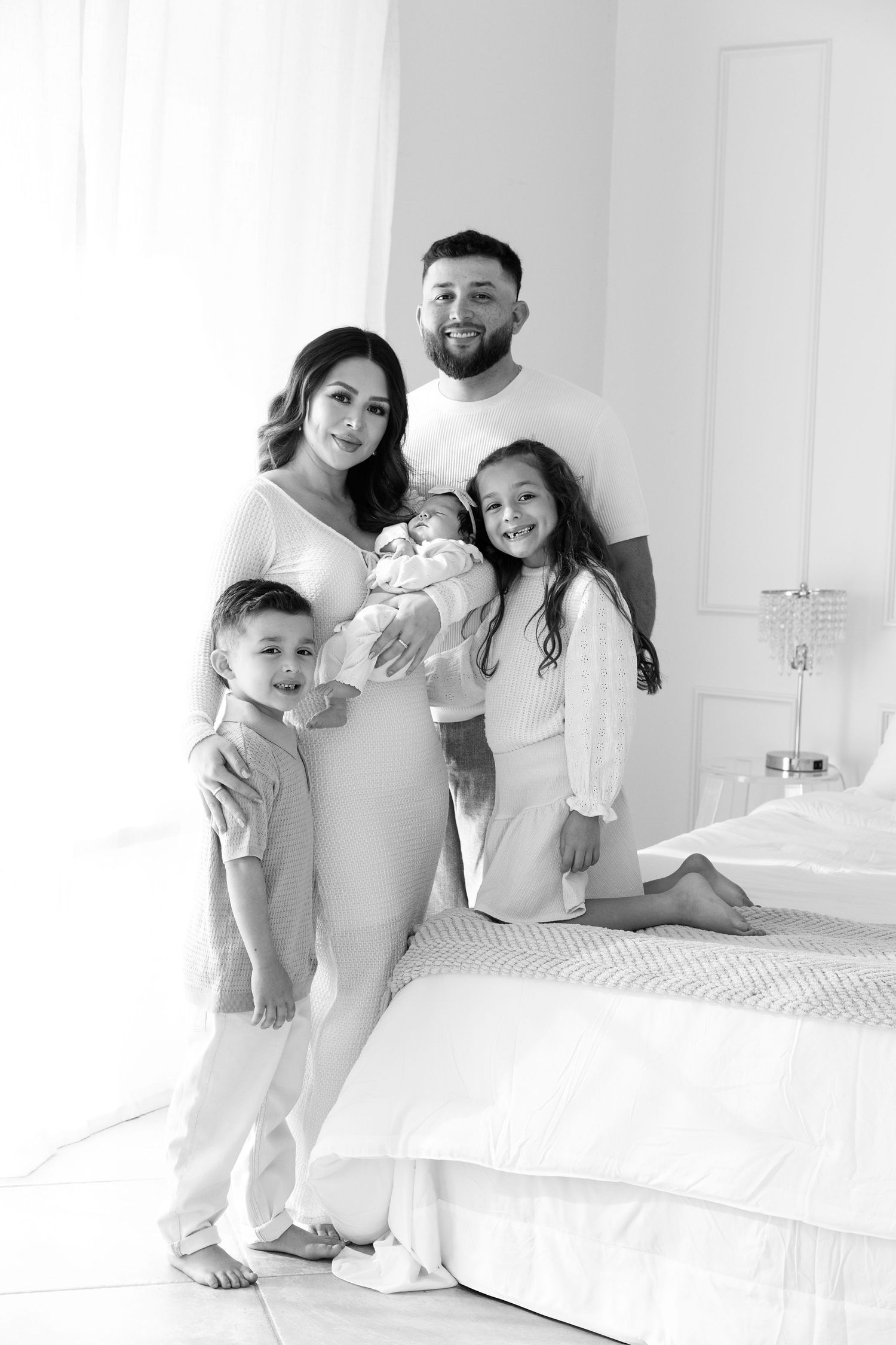 A family wearing white outfits poses together on a bed in a bright minimalist bedroom setting.