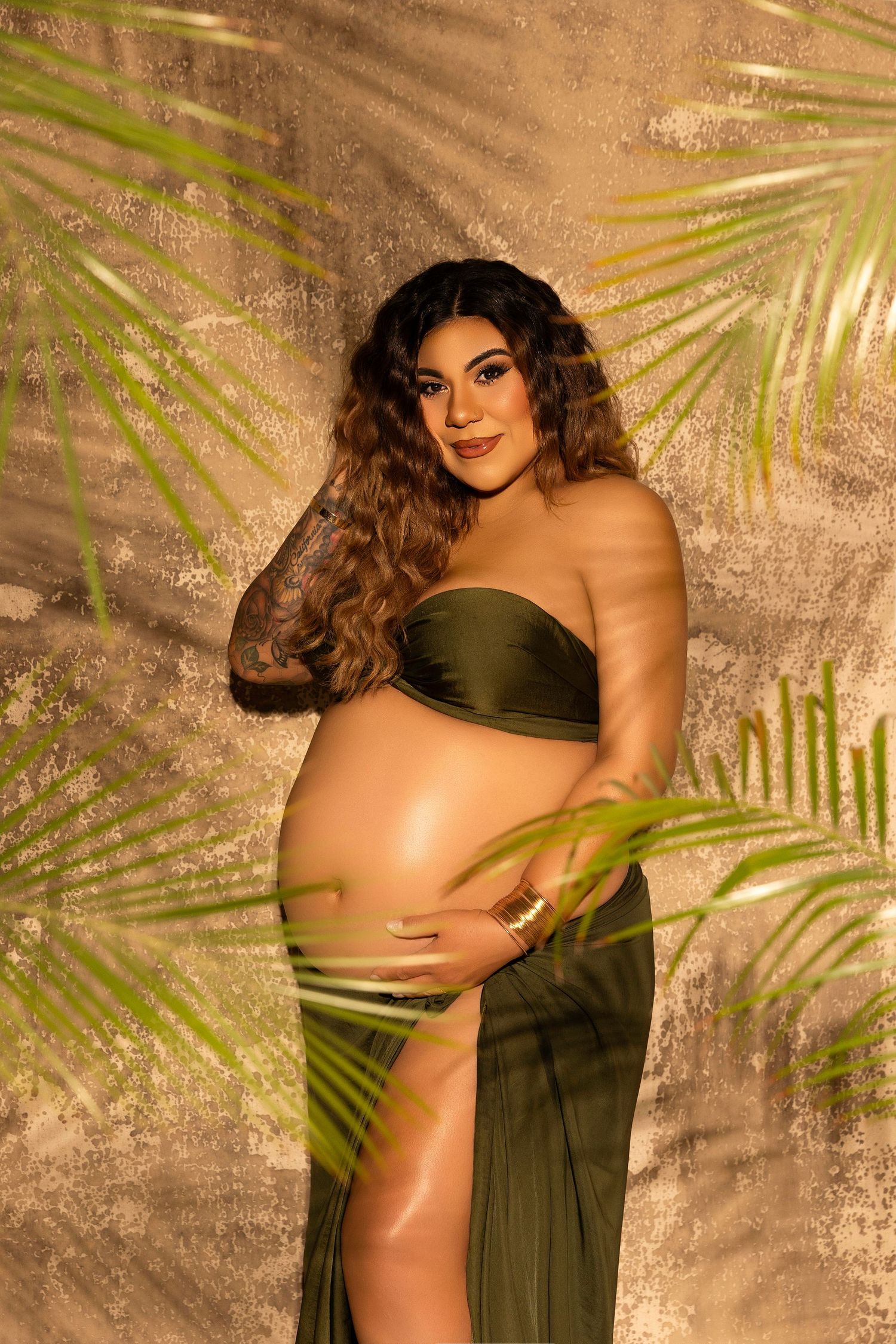 A maternity photo featuring an olive green dress against a tropical palm leaf backdrop with warm lighting.