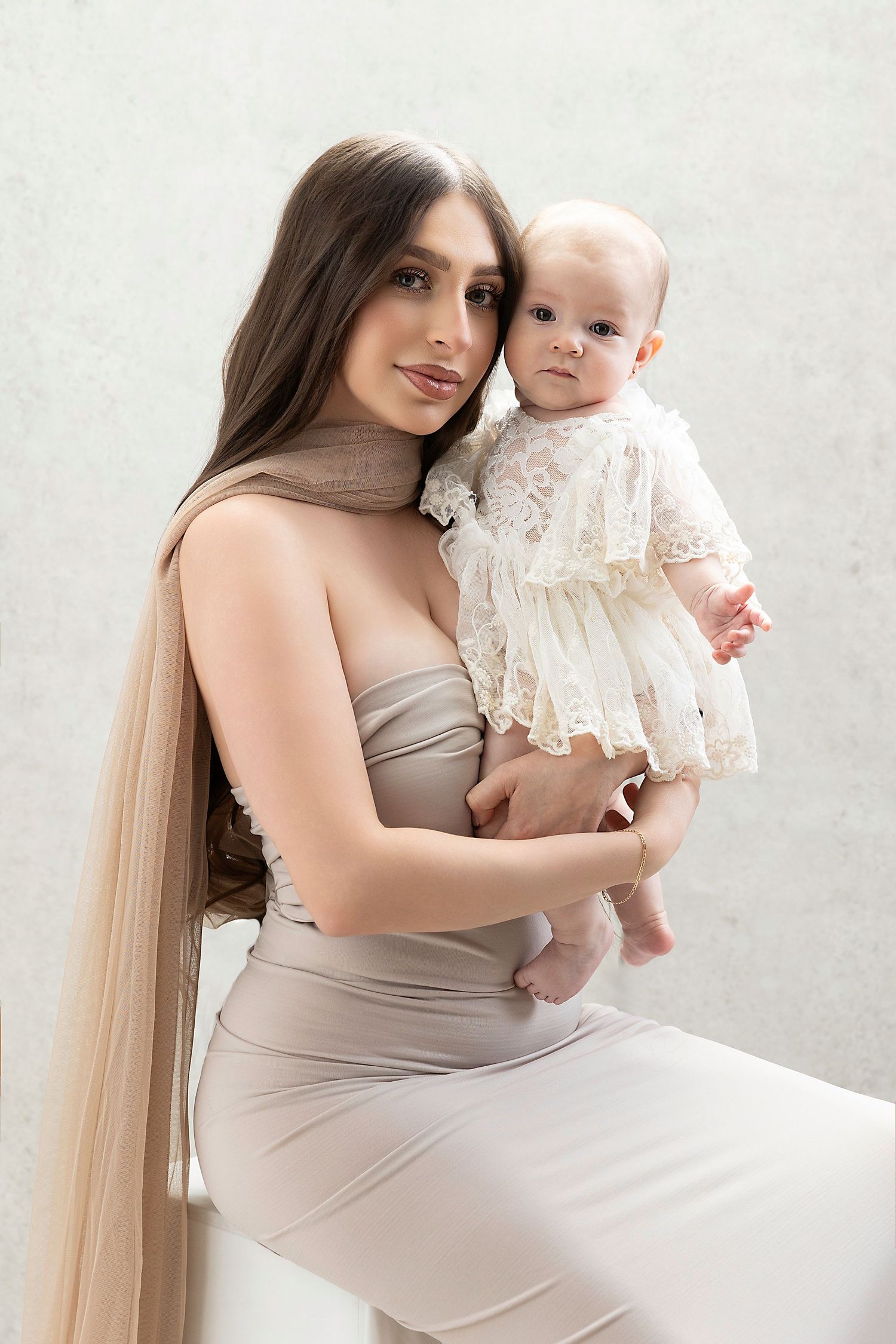 A parent in a beige dress holds a baby wearing a delicate white lace dress against a light background.