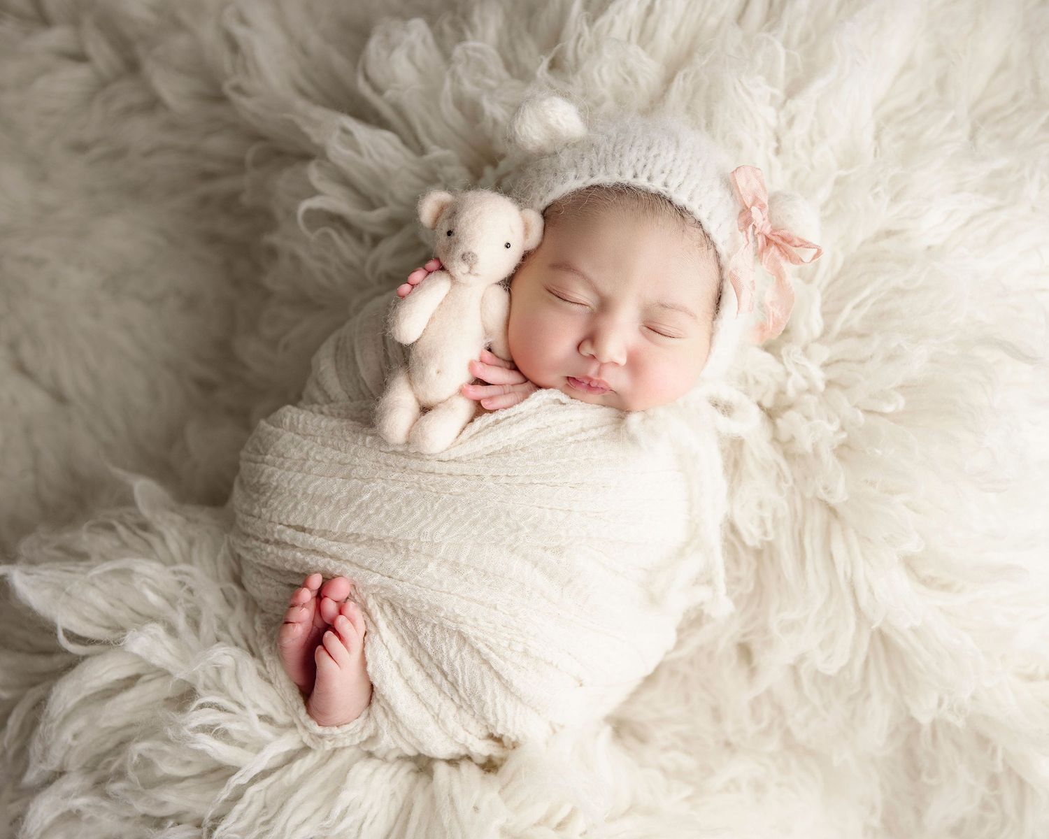 A peaceful sleeping newborn wrapped in white swaddle on fluffy fur blanket with small pink teddy bear.