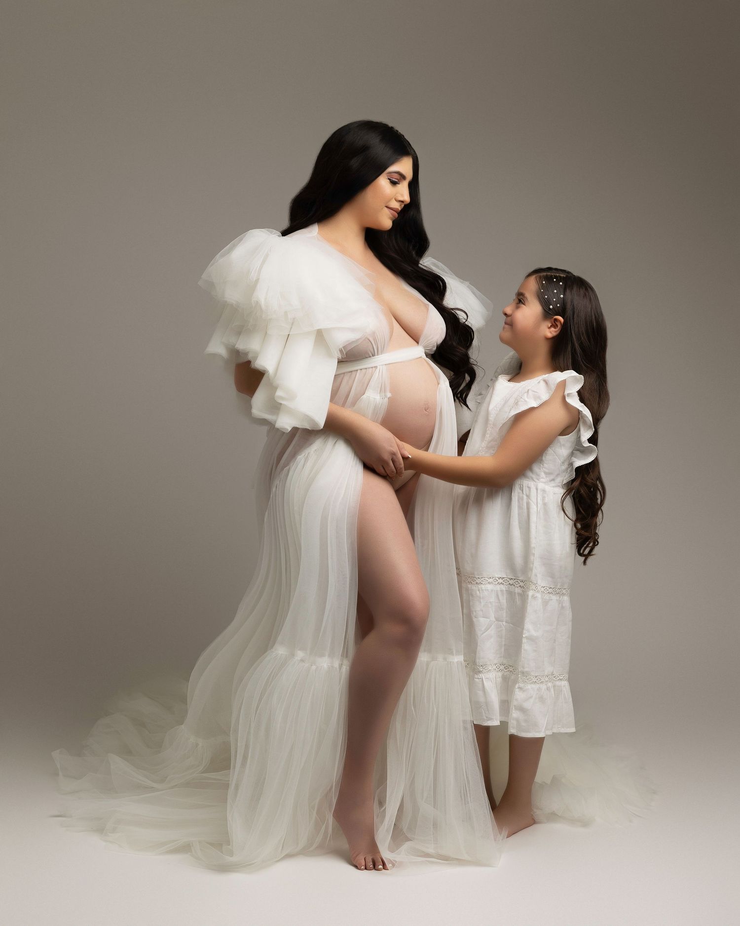 A tender maternity photoshoot shows two figures in flowing white dresses against a neutral backdrop.