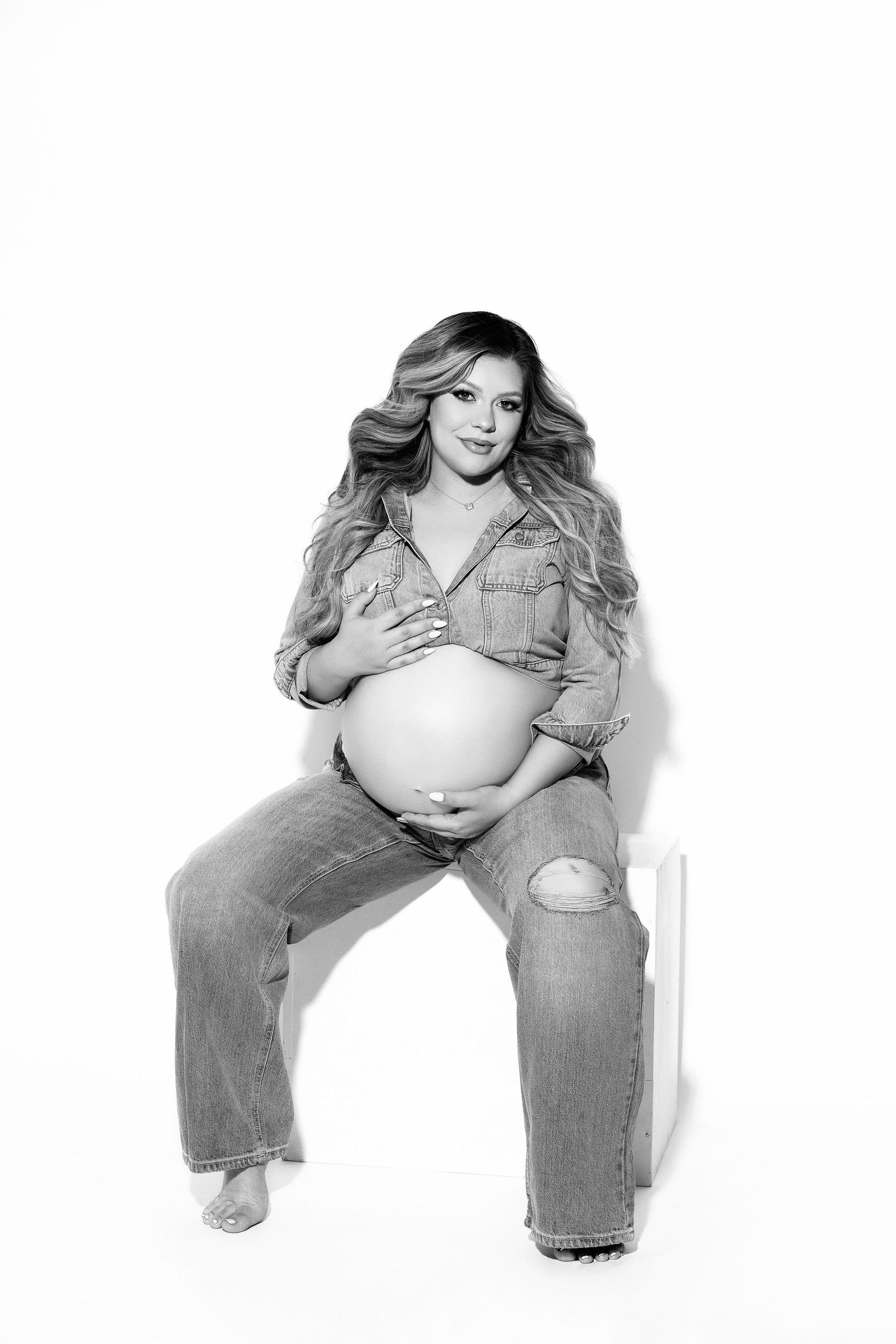 Black and white maternity photo of someone in casual denim outfit sitting against white background.