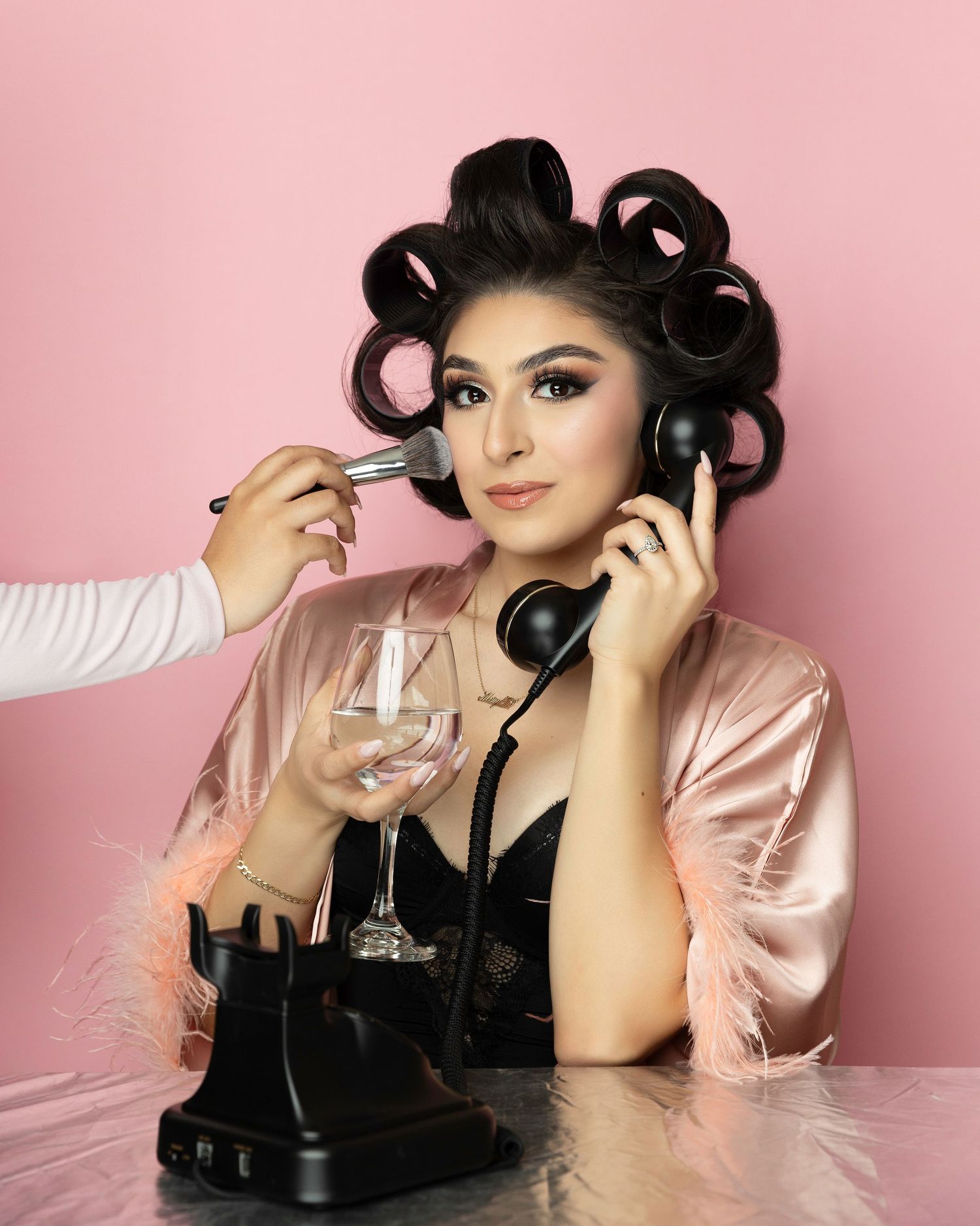 A beauty photoshoot against a pink backdrop featuring a model in a pink satin jacket and black dress.
