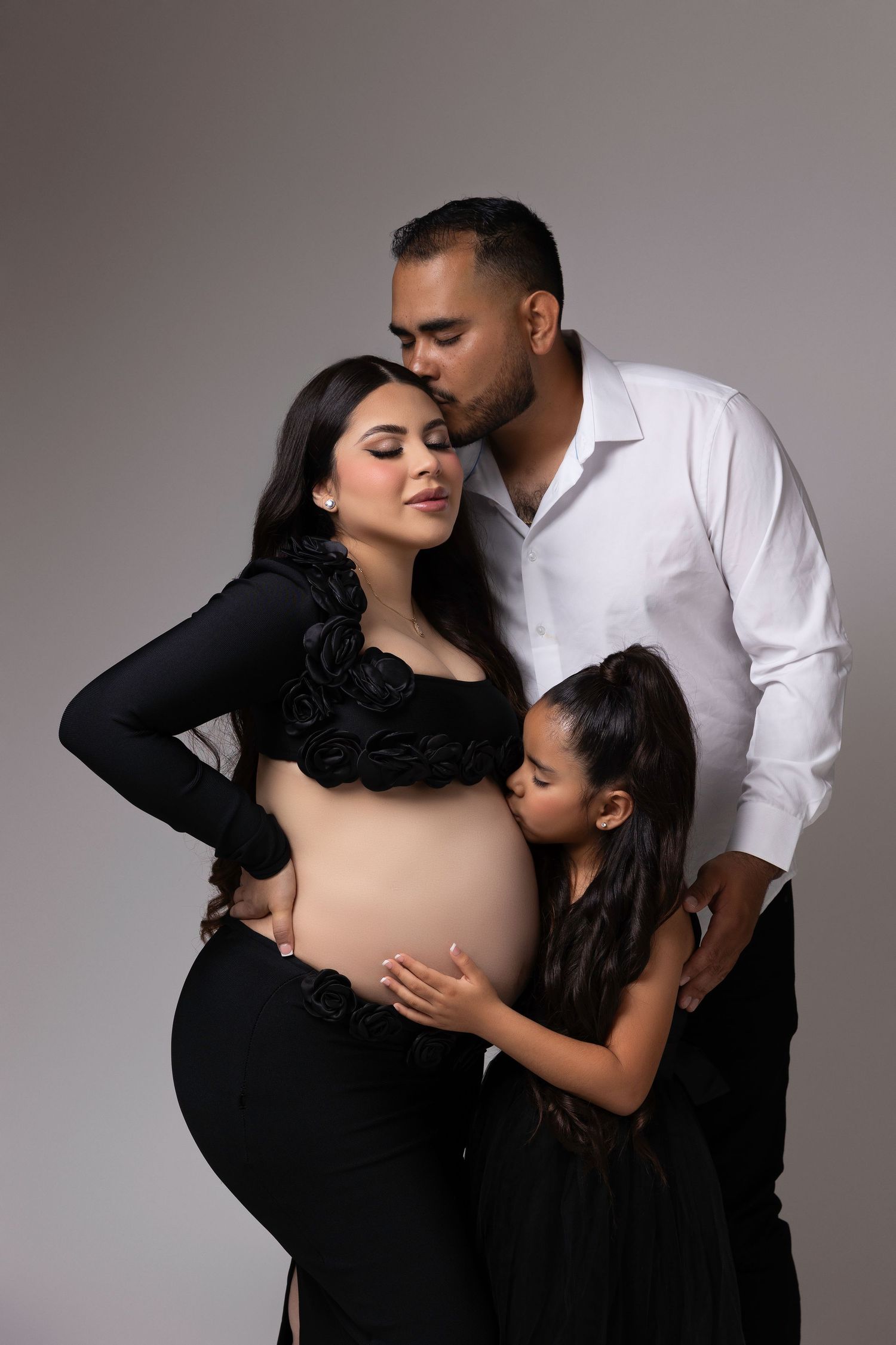 A romantic maternity photo series showing a couple in black and white outfits embracing in intimate poses against a gray backdrop.