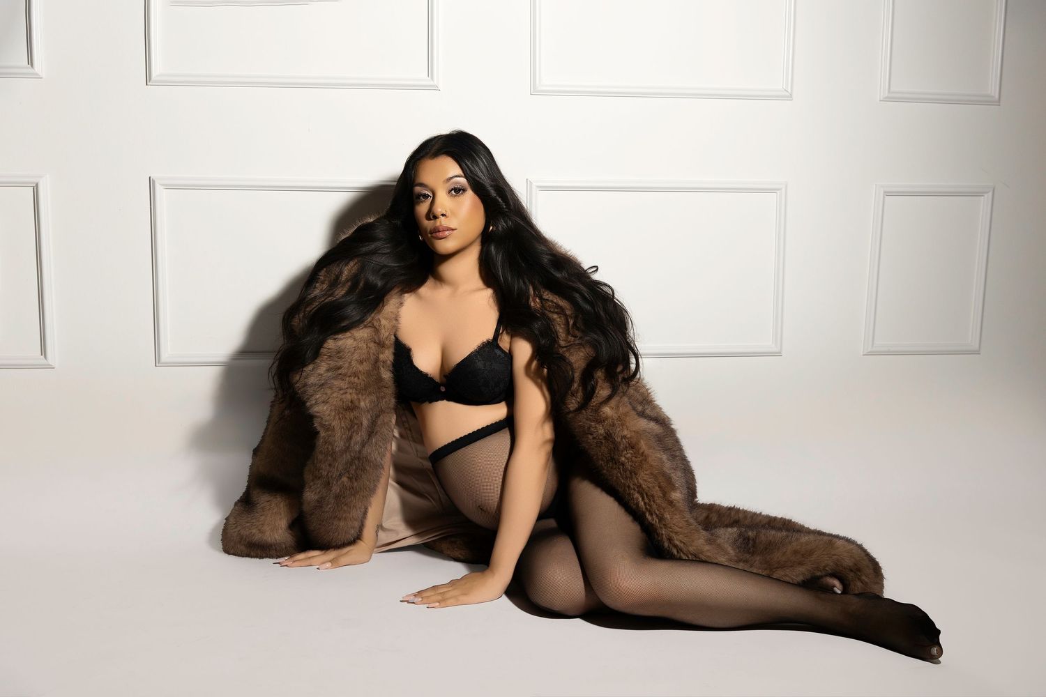 A figure in black sheer lingerie poses elegantly on a white backdrop with a brown fur coat.
