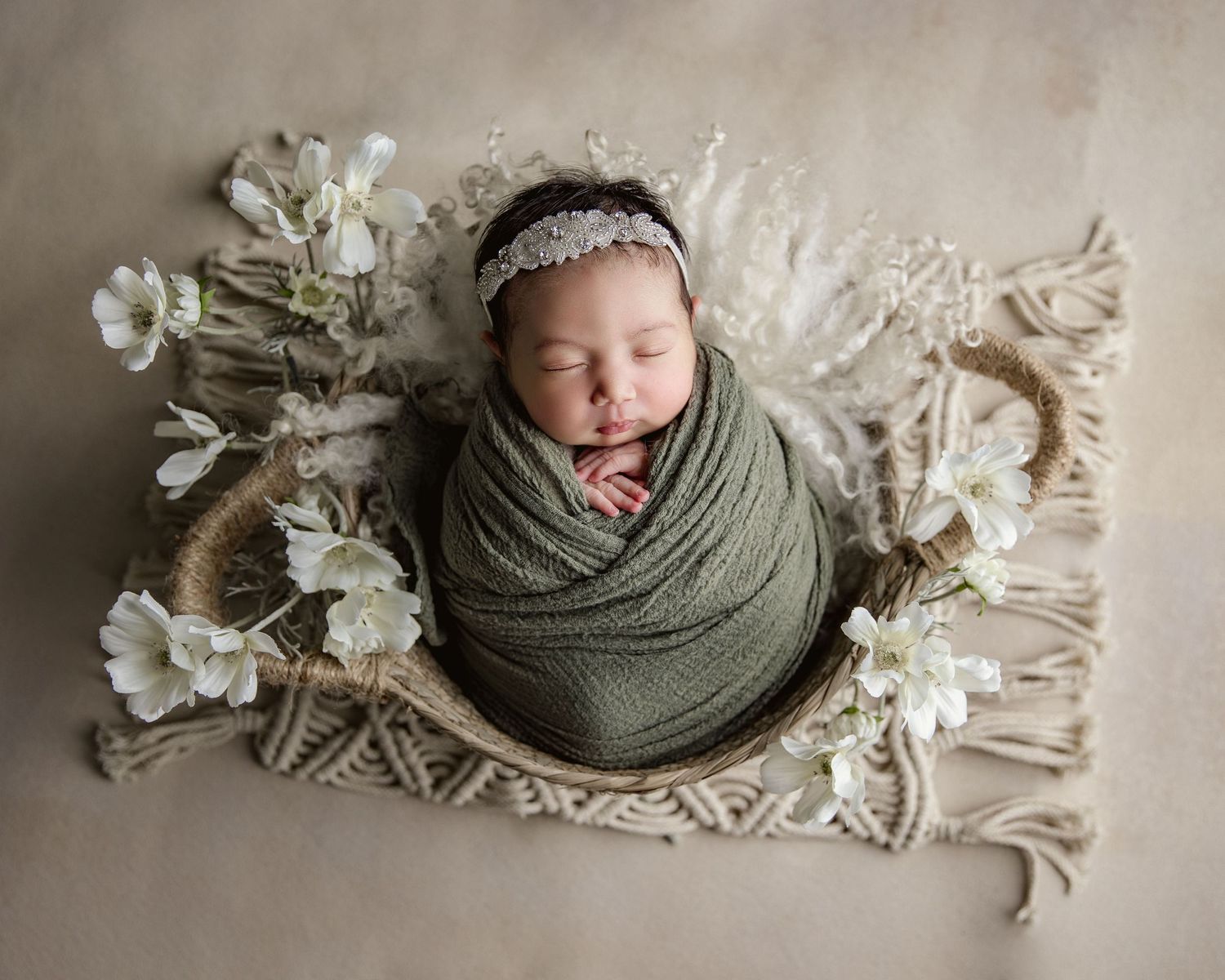 Newborn baby sleeping peacefully on cream blanket with sage green wrap and delicate white floral decorations.