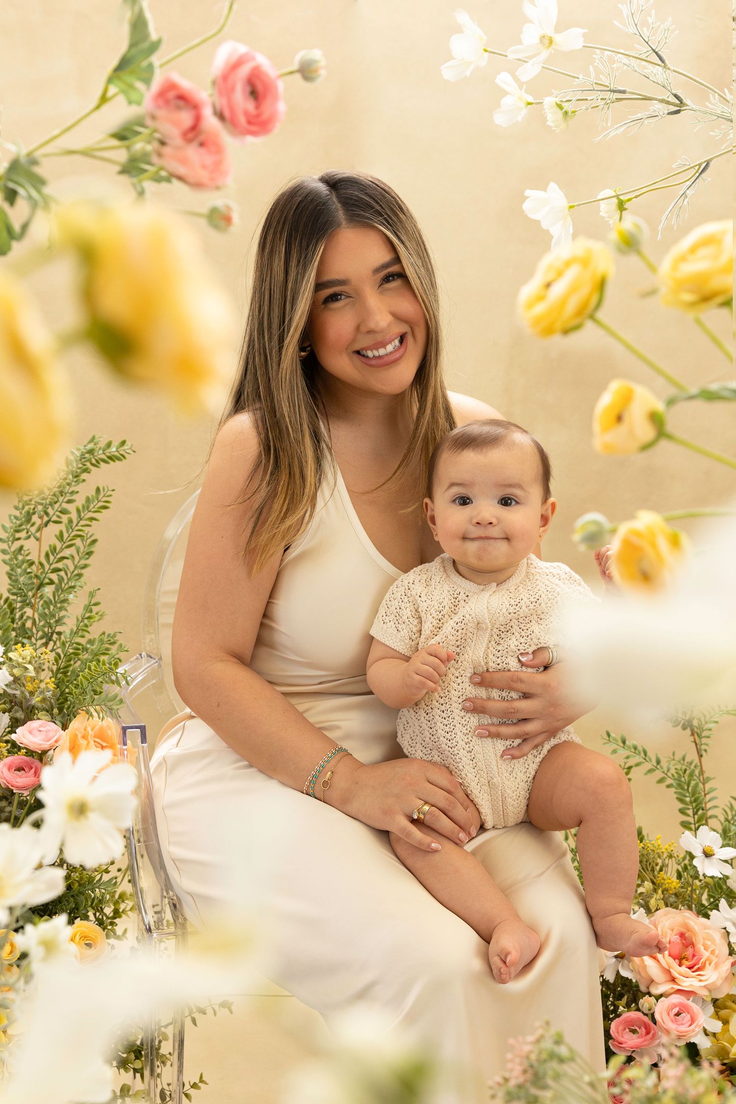 Mother and baby in white dresses pose together amid pink and yellow flowers in a dreamy spring garden setting.