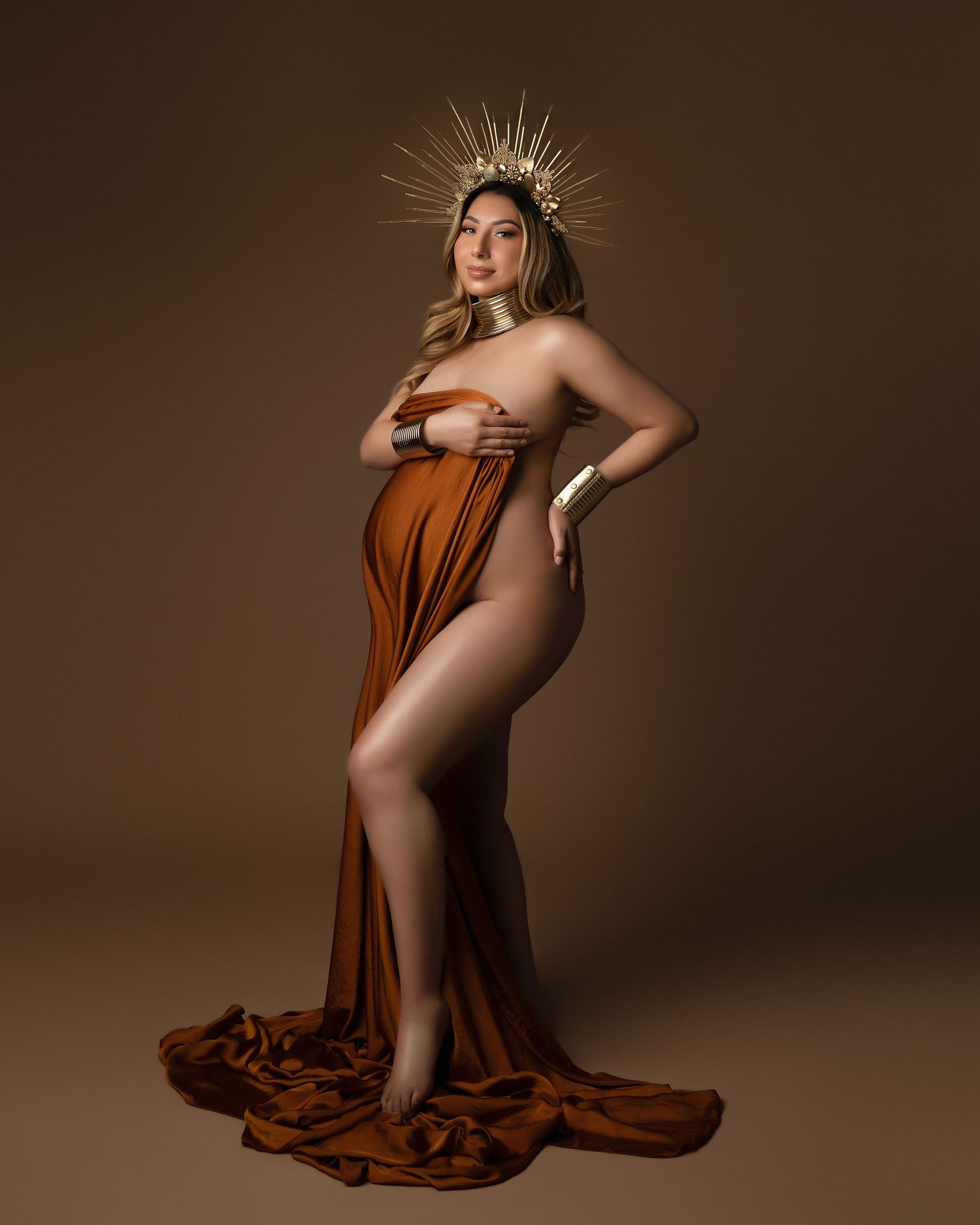 Pregnant woman in rust orange draped fabric and gold crown poses gracefully against a warm brown studio background.