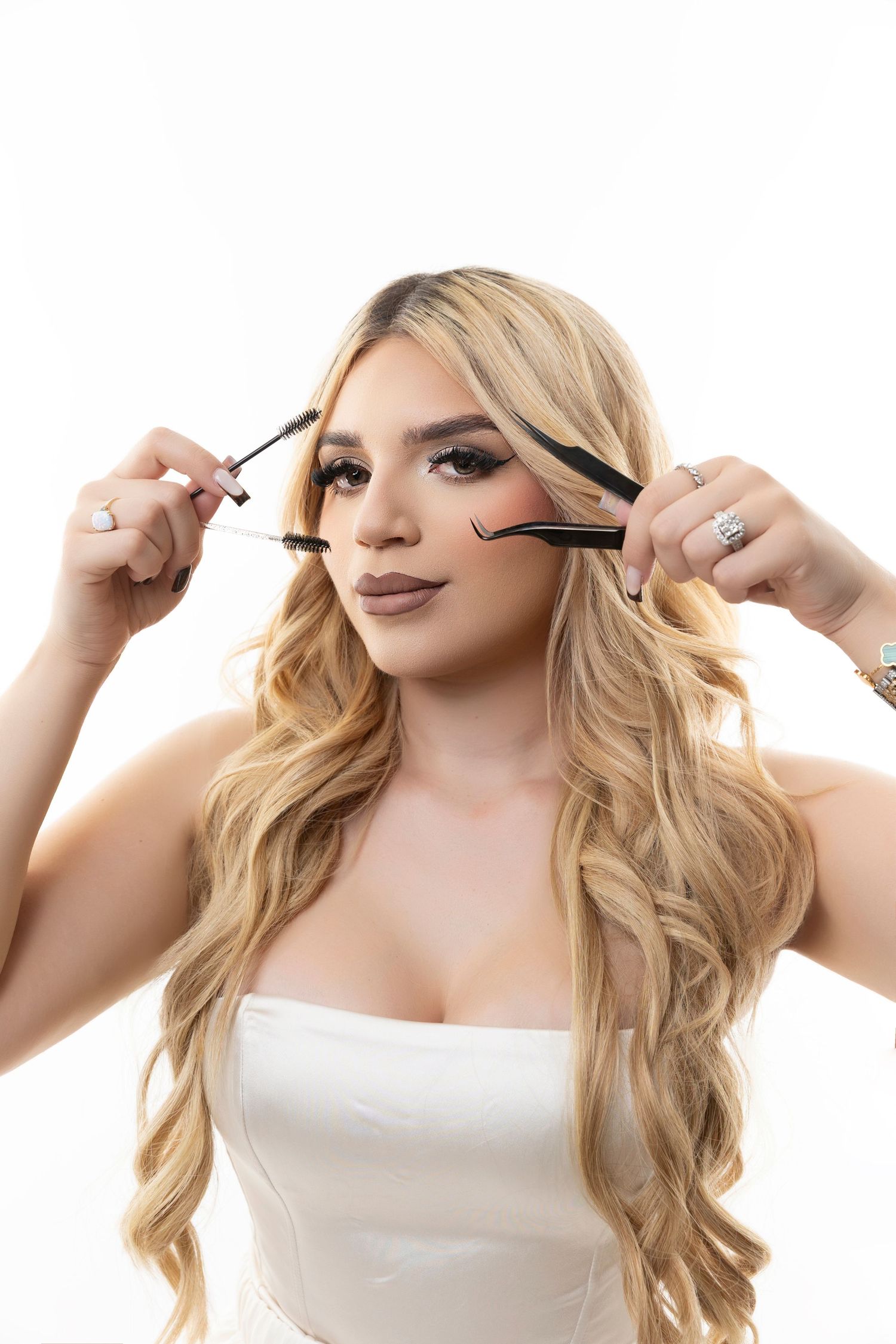 A person with long blonde wavy hair demonstrates applying mascara in a white top against a white background.