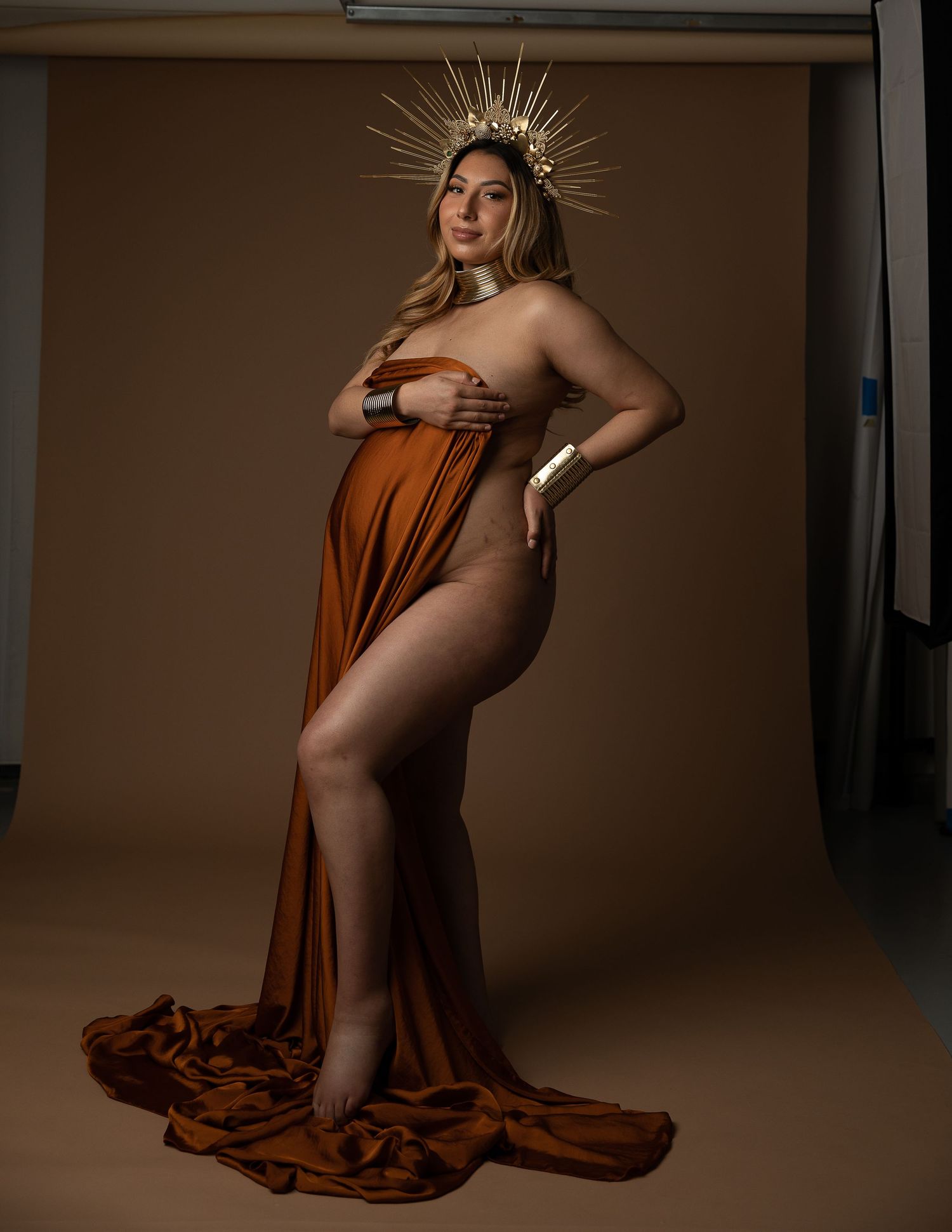 Pregnant woman in rust orange draped fabric gown wearing gold sun crown poses elegantly in professional maternity photo shoot.