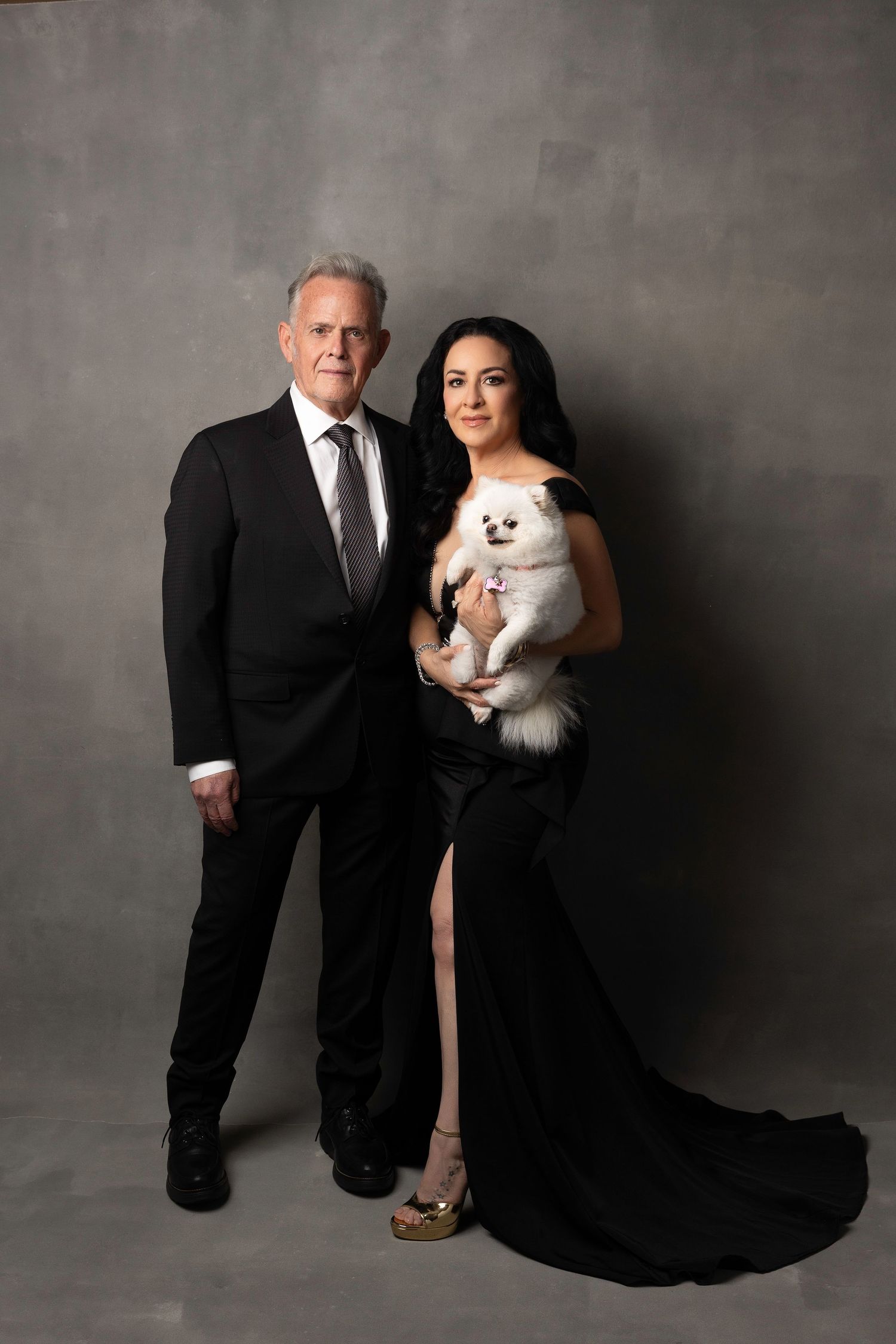 A couple in formal black attire poses with a fluffy white Pomeranian dog against a dark studio backdrop.
