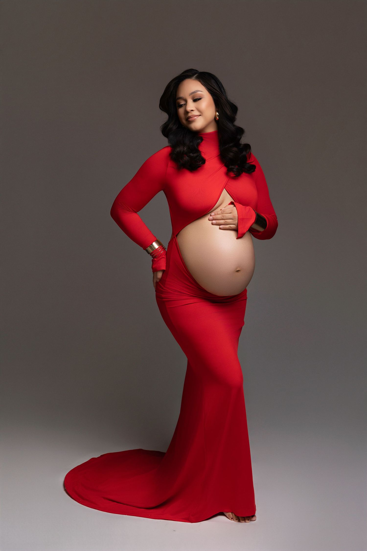 A pregnant woman in a long-sleeved red dress poses elegantly against a grey studio backdrop for a maternity photoshoot.
