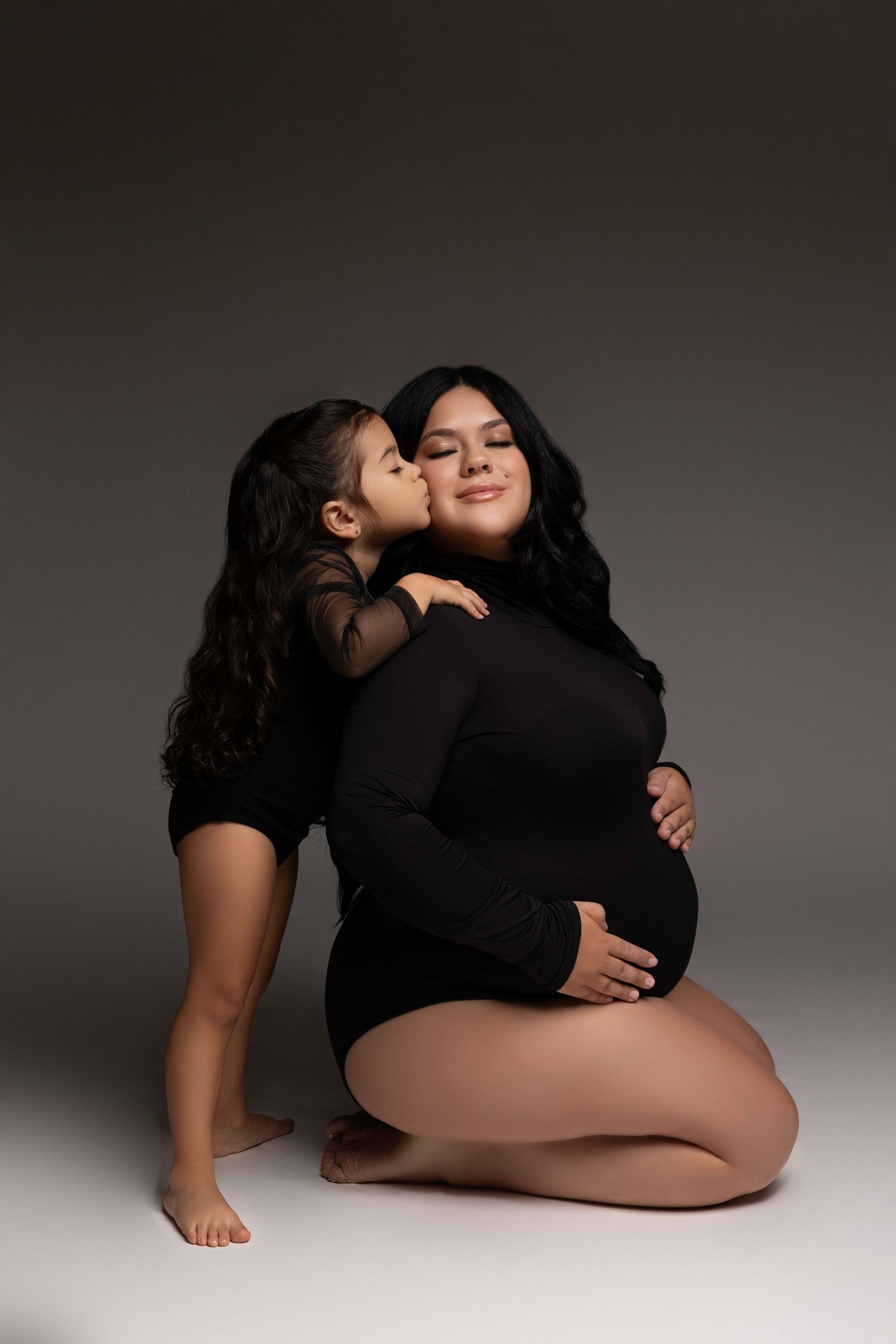 An intimate maternity portrait of two figures in black clothing embracing against a dark background.