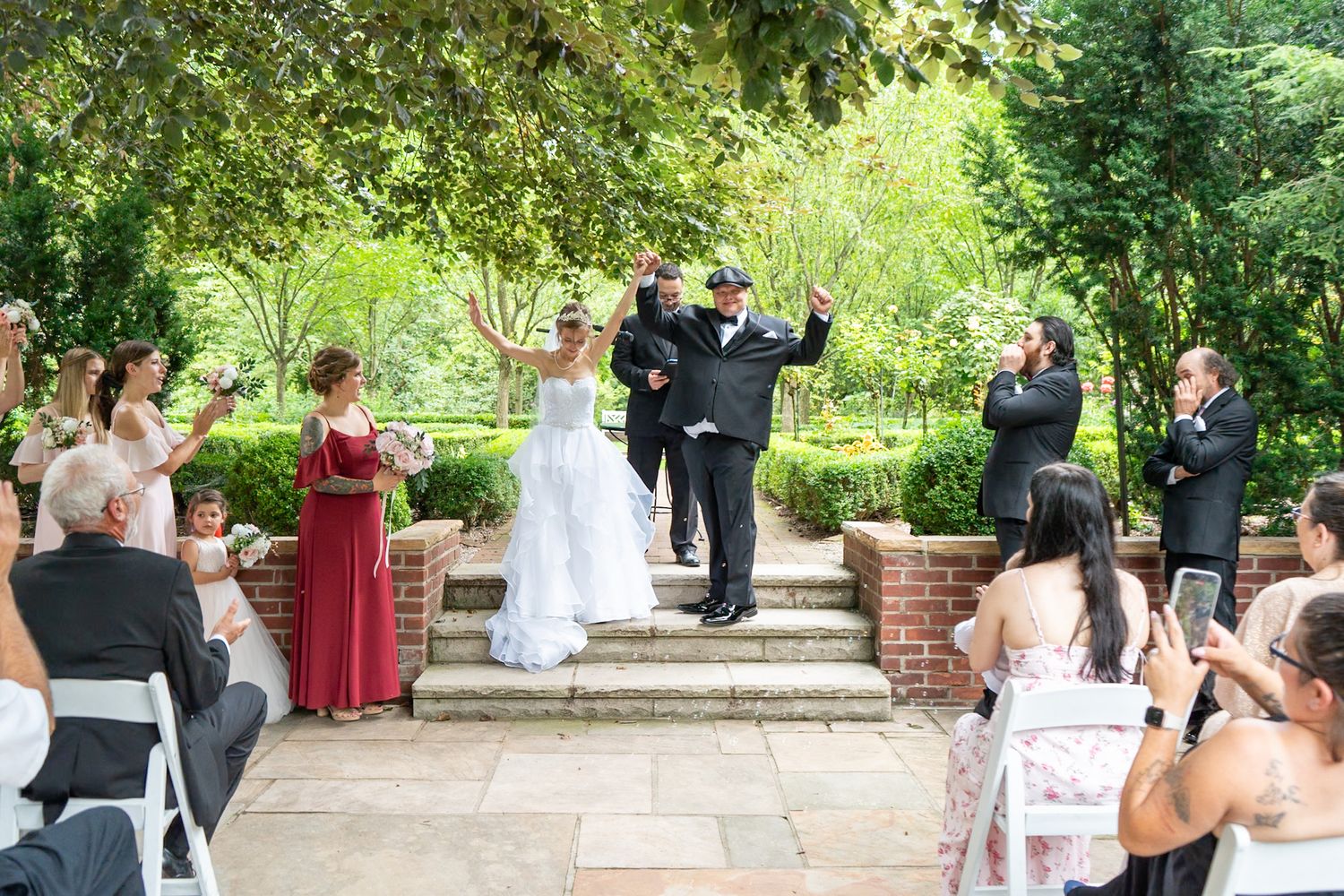 Bride and groom raise their arms in celebration on garden steps as wedding guests cheer and photograph the outdoor ceremony.