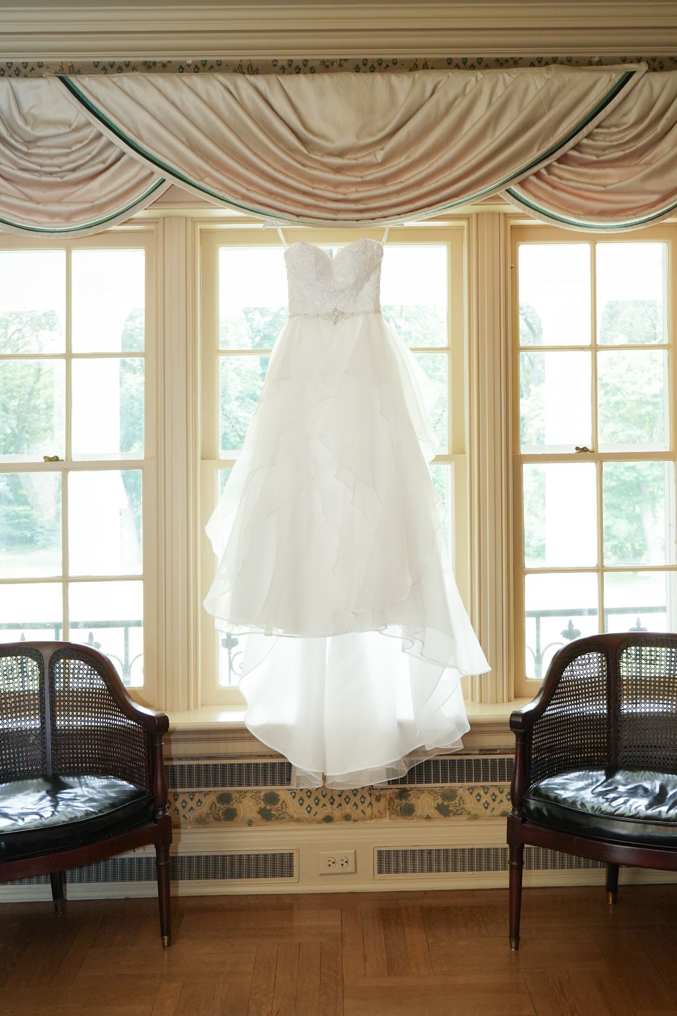 White strapless wedding gown with layered tulle skirt hanging in a bright window with silk swag valance between two wicker chairs.