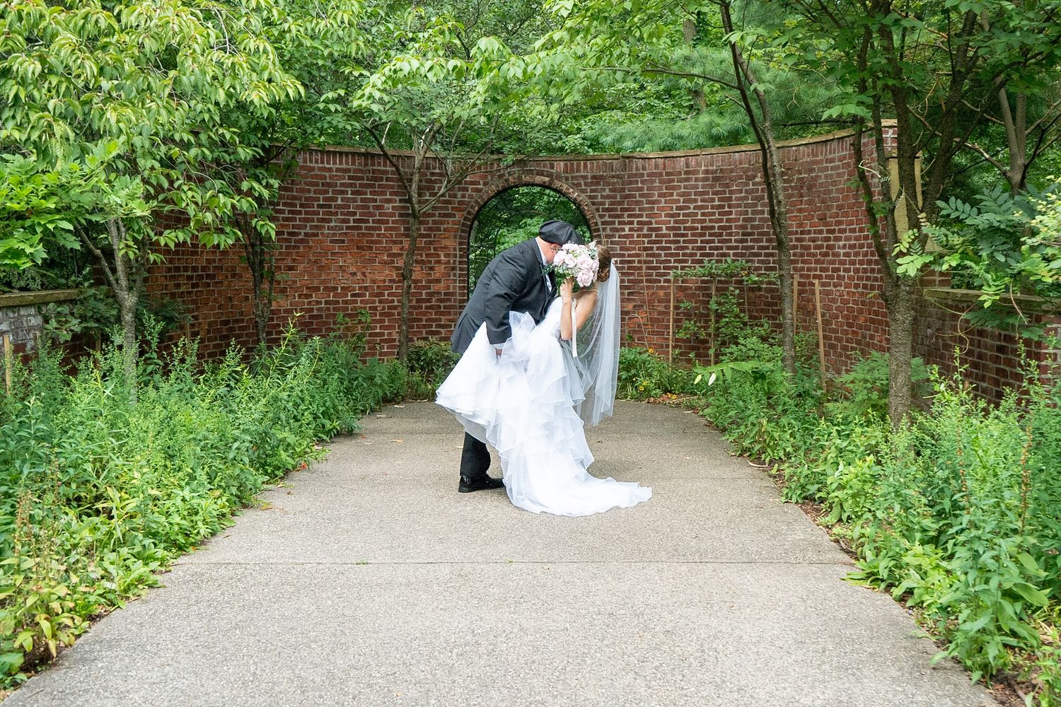 Bride and groom share a romantic dip kiss on a garden path in front of a historic brick wall with an arched opening and lush greenery.