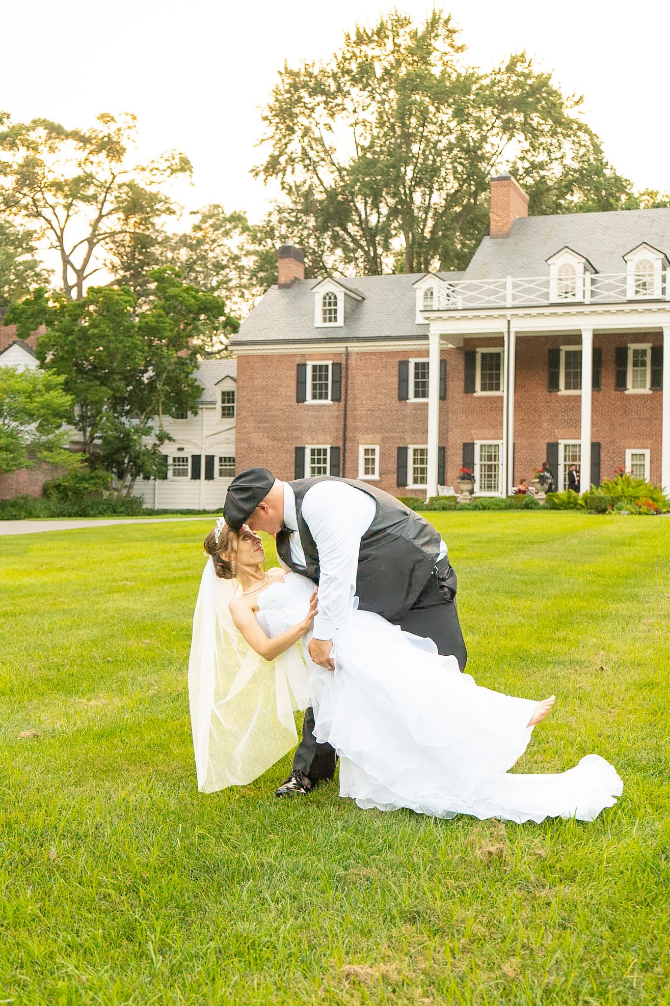 Groom in vest and cap dips bride in a white gown on a sunny lawn in front of a red brick Georgian mansion wedding venue.