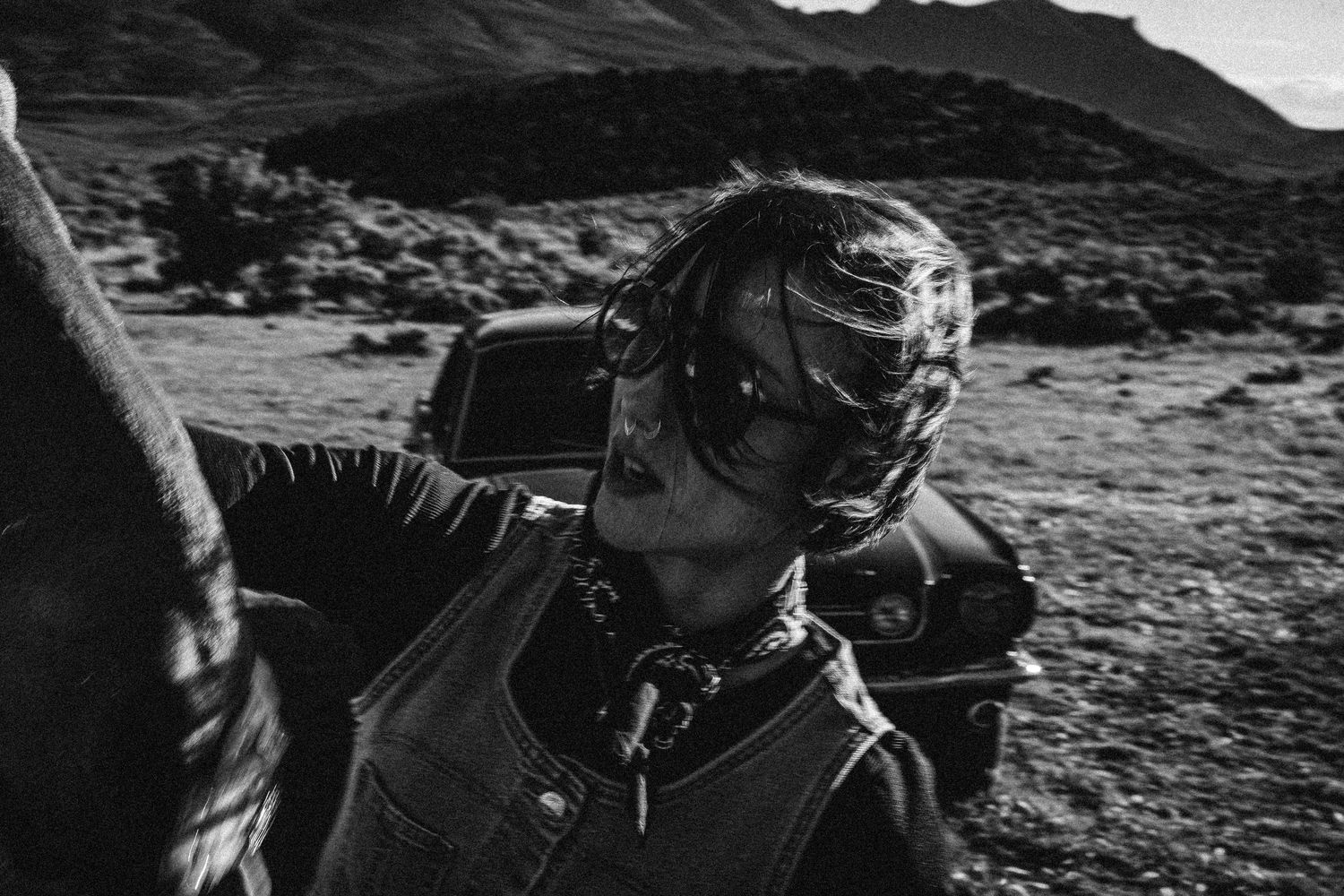 Black and white photo of a woman in sunglasses and leather jacket on a motorcycle in a desert landscape.
