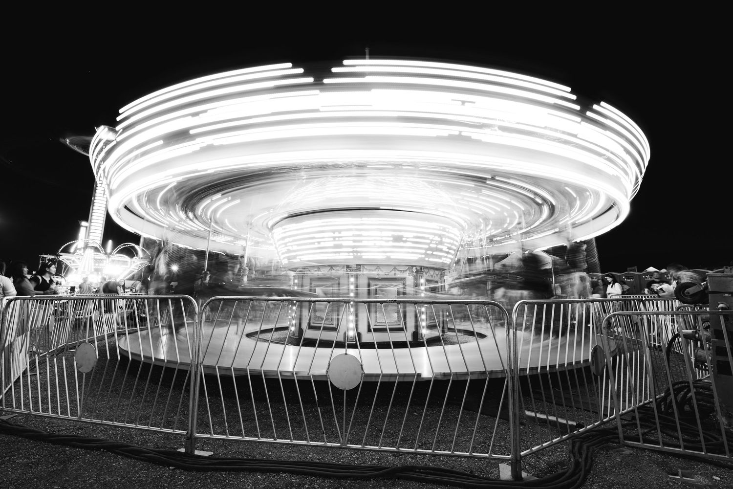 Long exposure black and white photo of a spinning carousel at night with motion blur light trails.