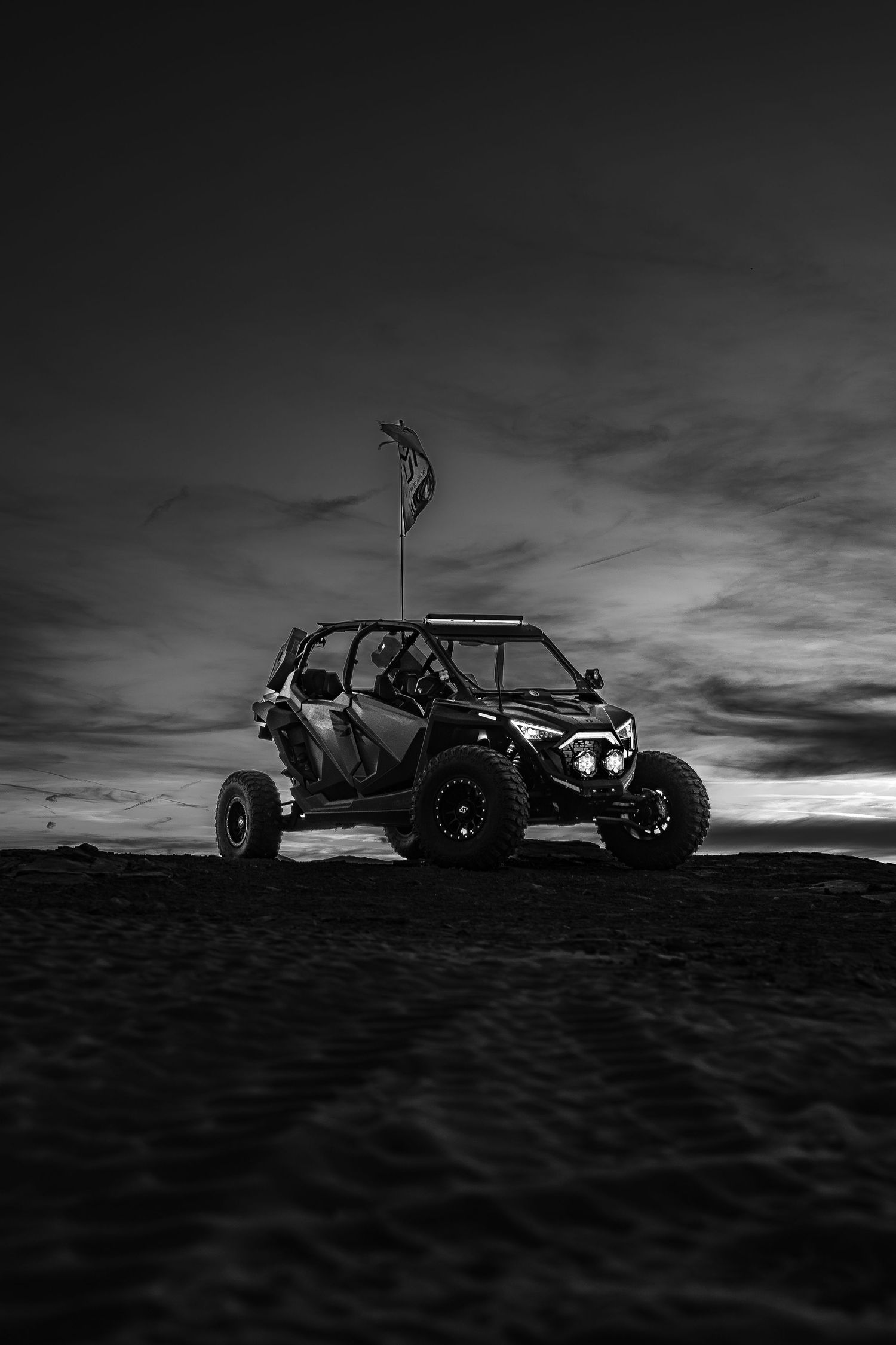 Black and white UTV parked on dark desert terrain under dramatic overcast sky with flag.
