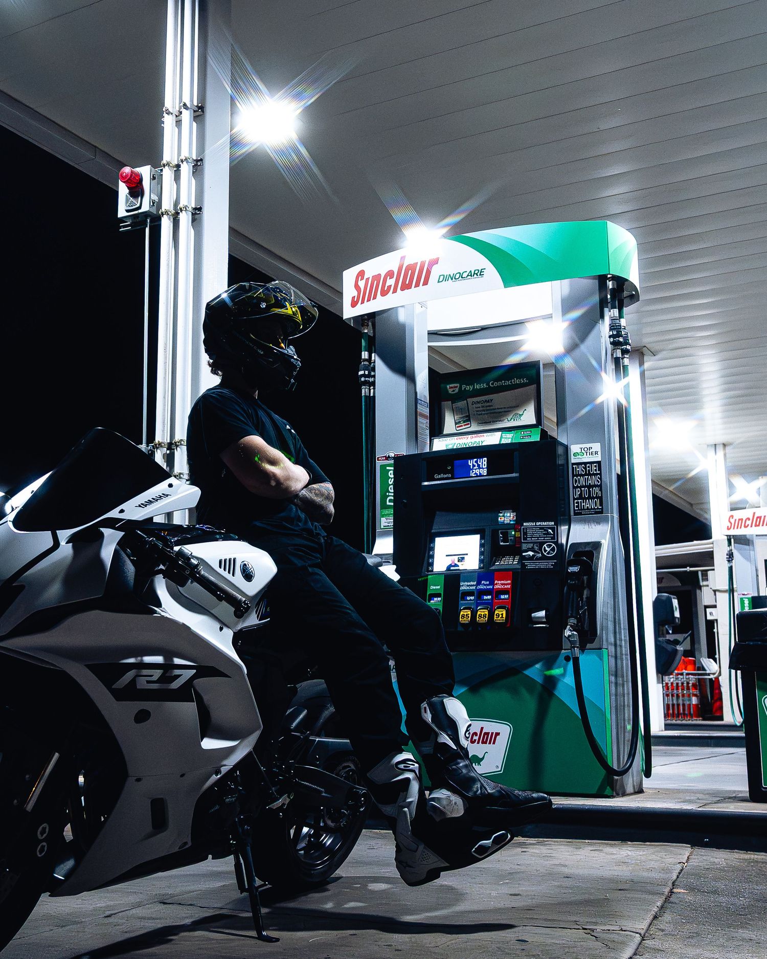 Helmeted rider in all-black gear leans against white Yamaha R7 at Sinclair gas station at night.