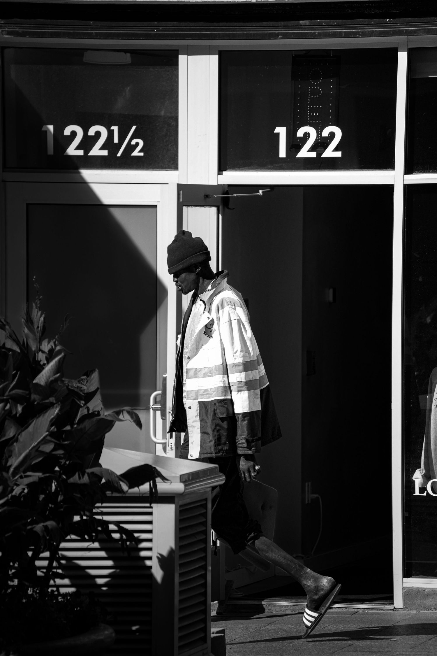 A person in a reflective safety vest walks past building entrances numbered 122 and 122½ in black and white.