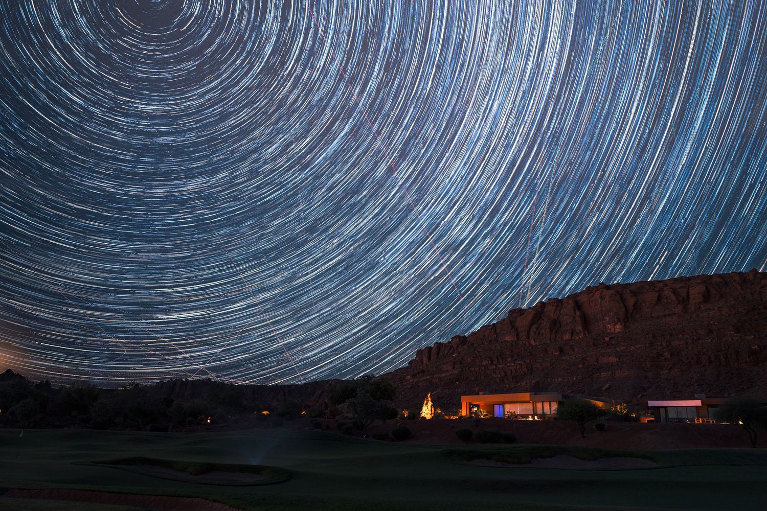 Long exposure star trail photography over a desert landscape with a glowing building below red cliffs.