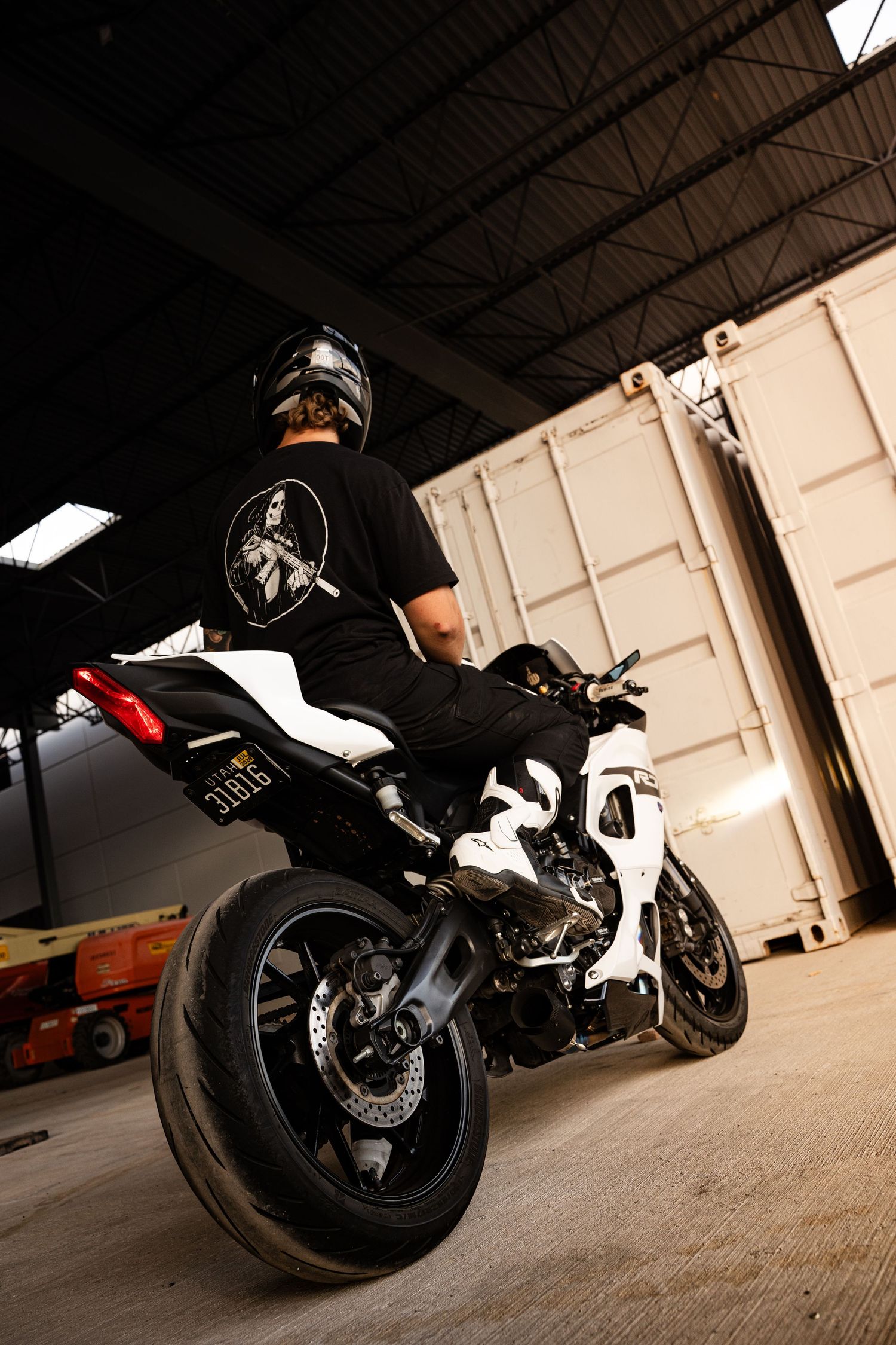 Rear view of a motorcyclist in black gear and helmet seated on a white sport motorcycle inside an industrial warehouse.