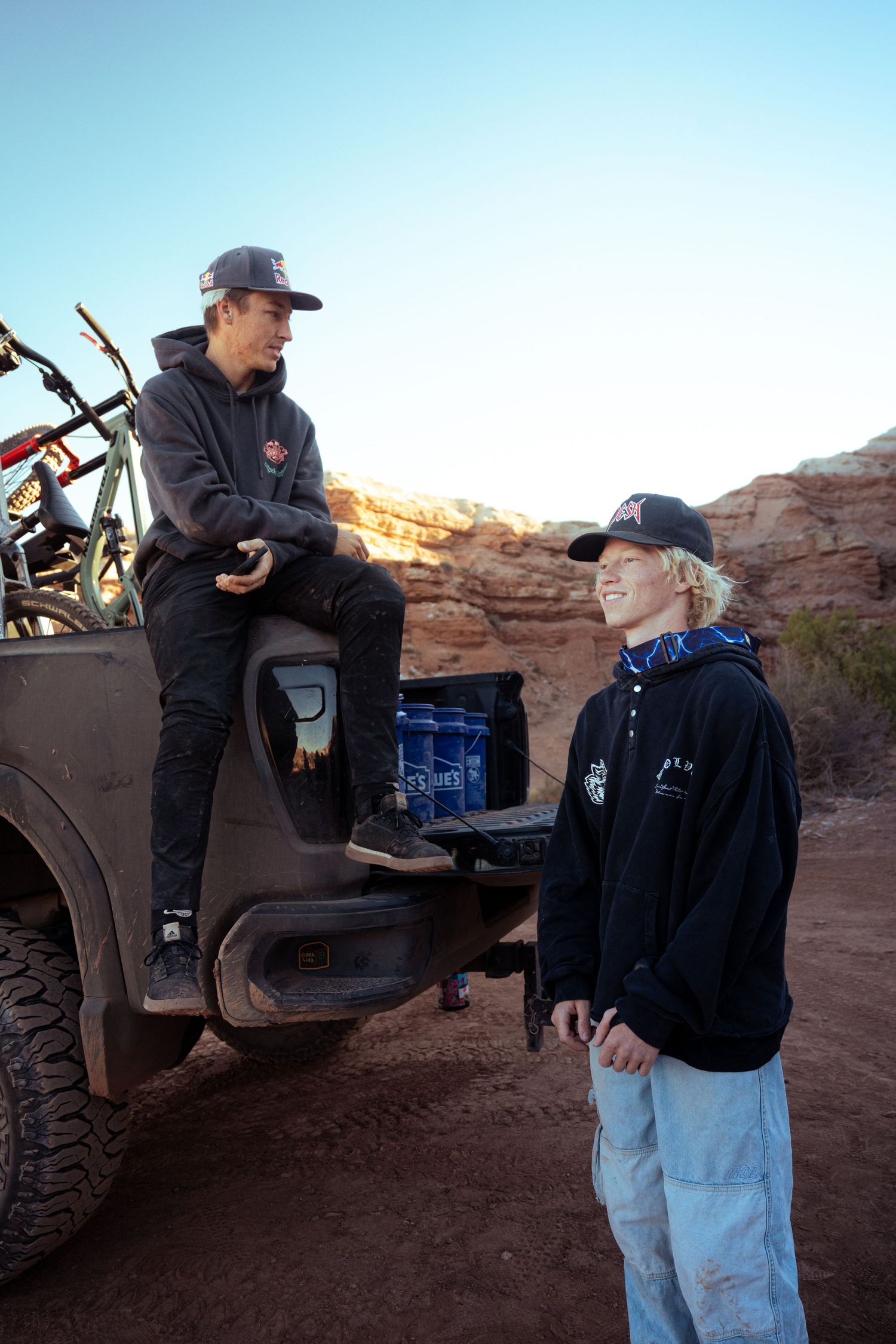 Two young mountain bikers chat near truck tailgate with bikes in red rock desert landscape.