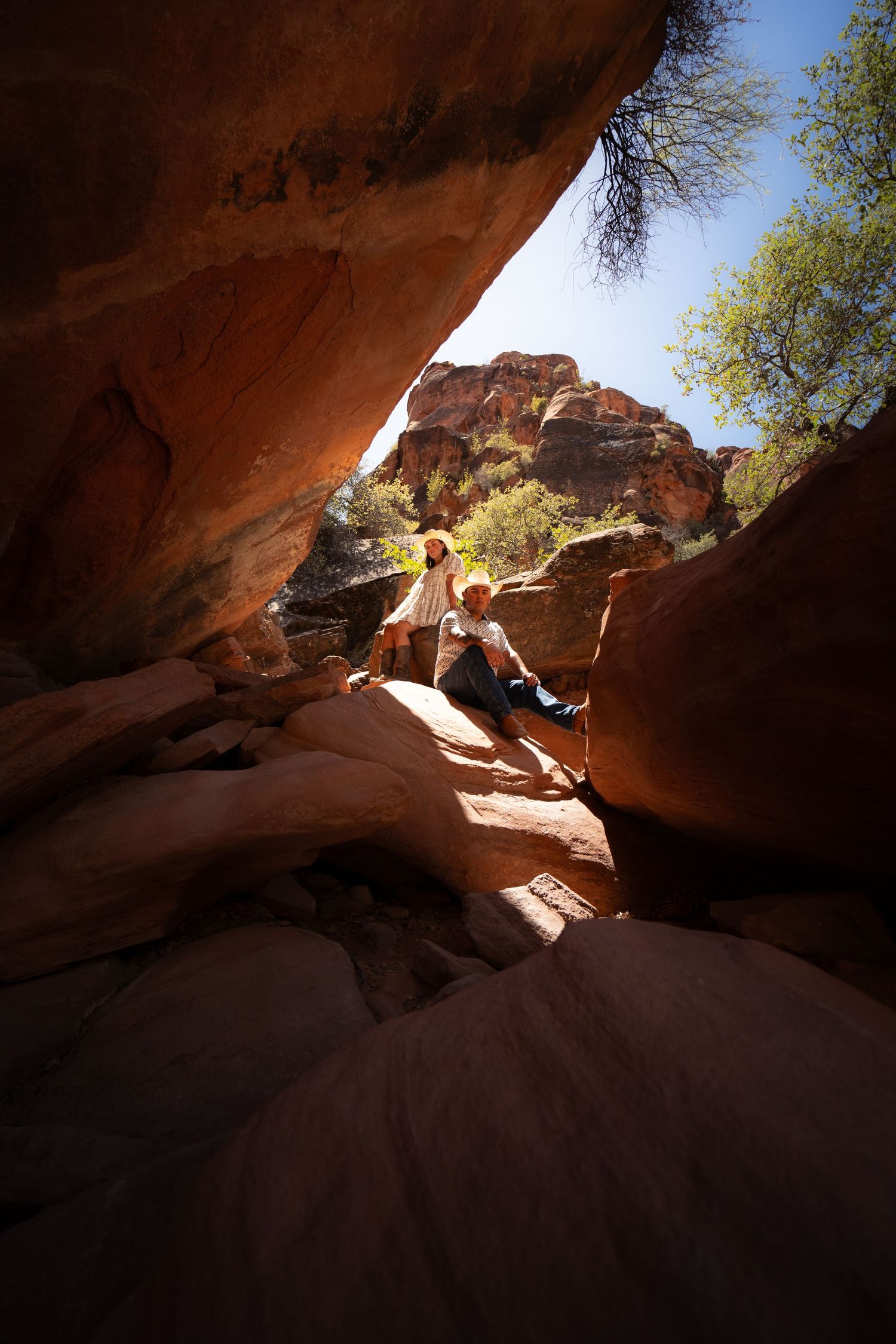 Two hikers explore a dramatic red rock canyon formation under a bright blue sky in a desert landscape.
