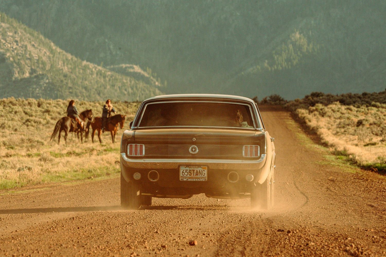 Rear view of a classic Ford Mustang driving down a dusty dirt road toward a group of horseback riders.