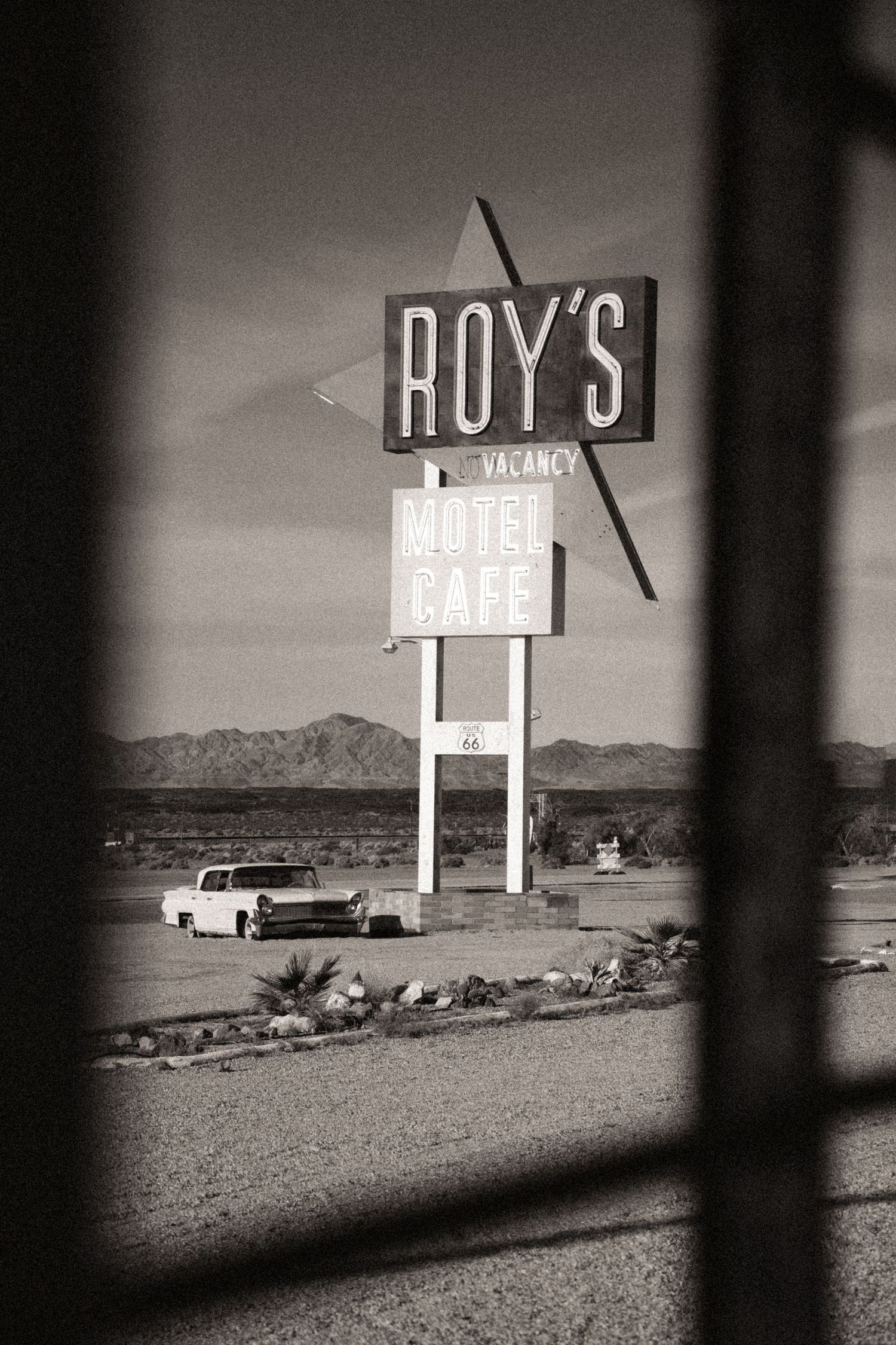 Black and white photo of Roy's Motel Cafe sign on Route 66 with a vintage car parked in the desert landscape.