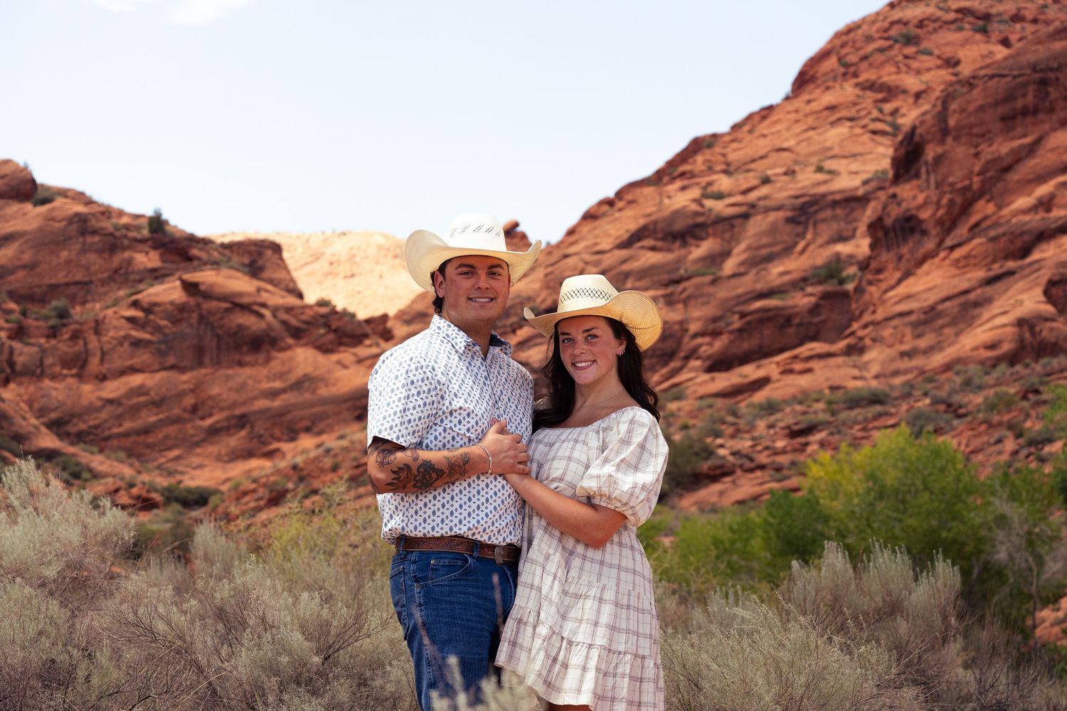 Smiling couple in cowboy hats embracing before stunning red rock canyon landscape.